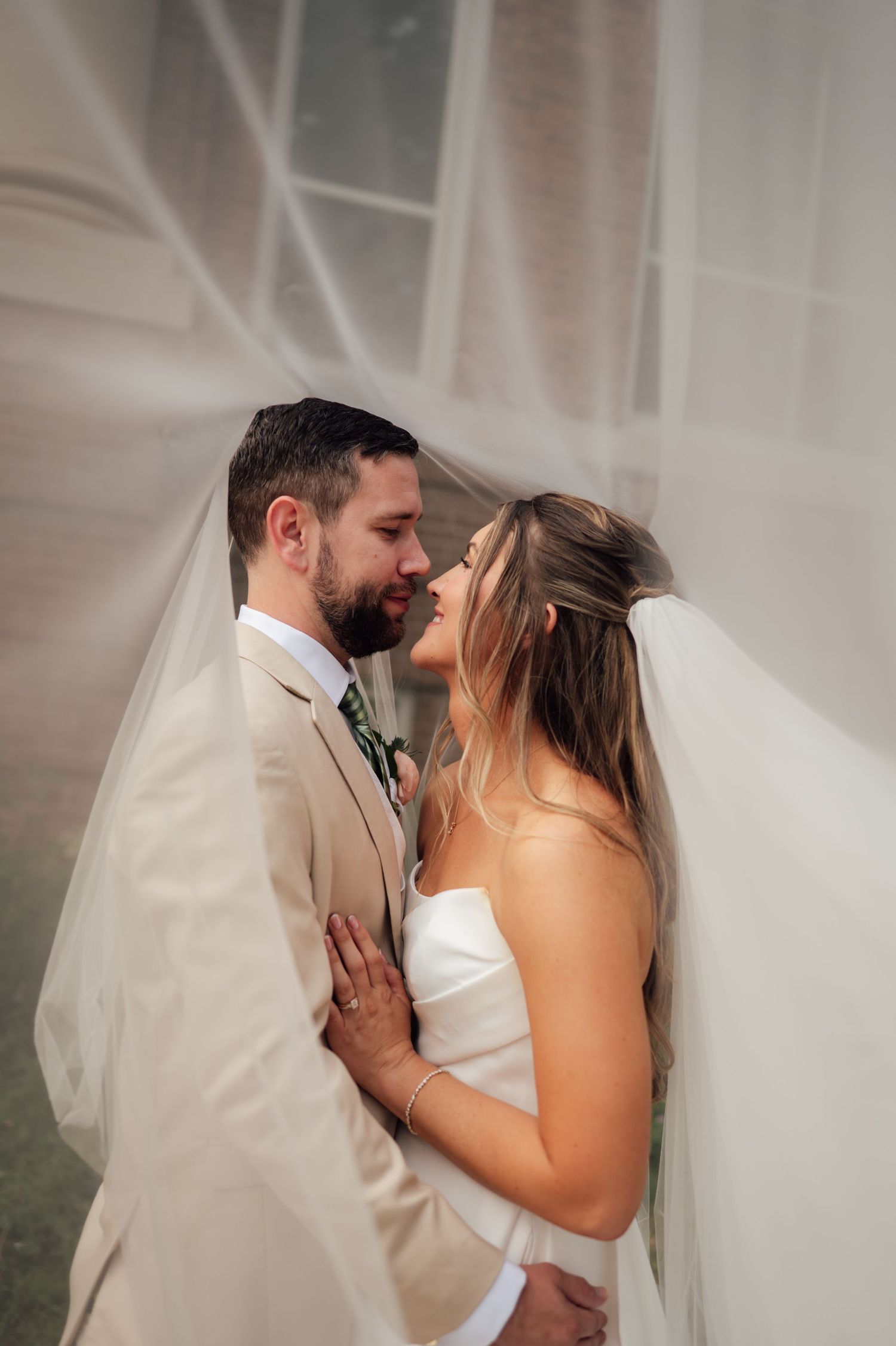 Bride and groom share a romantic moment under a flowing white veil, dressed in wedding attire.