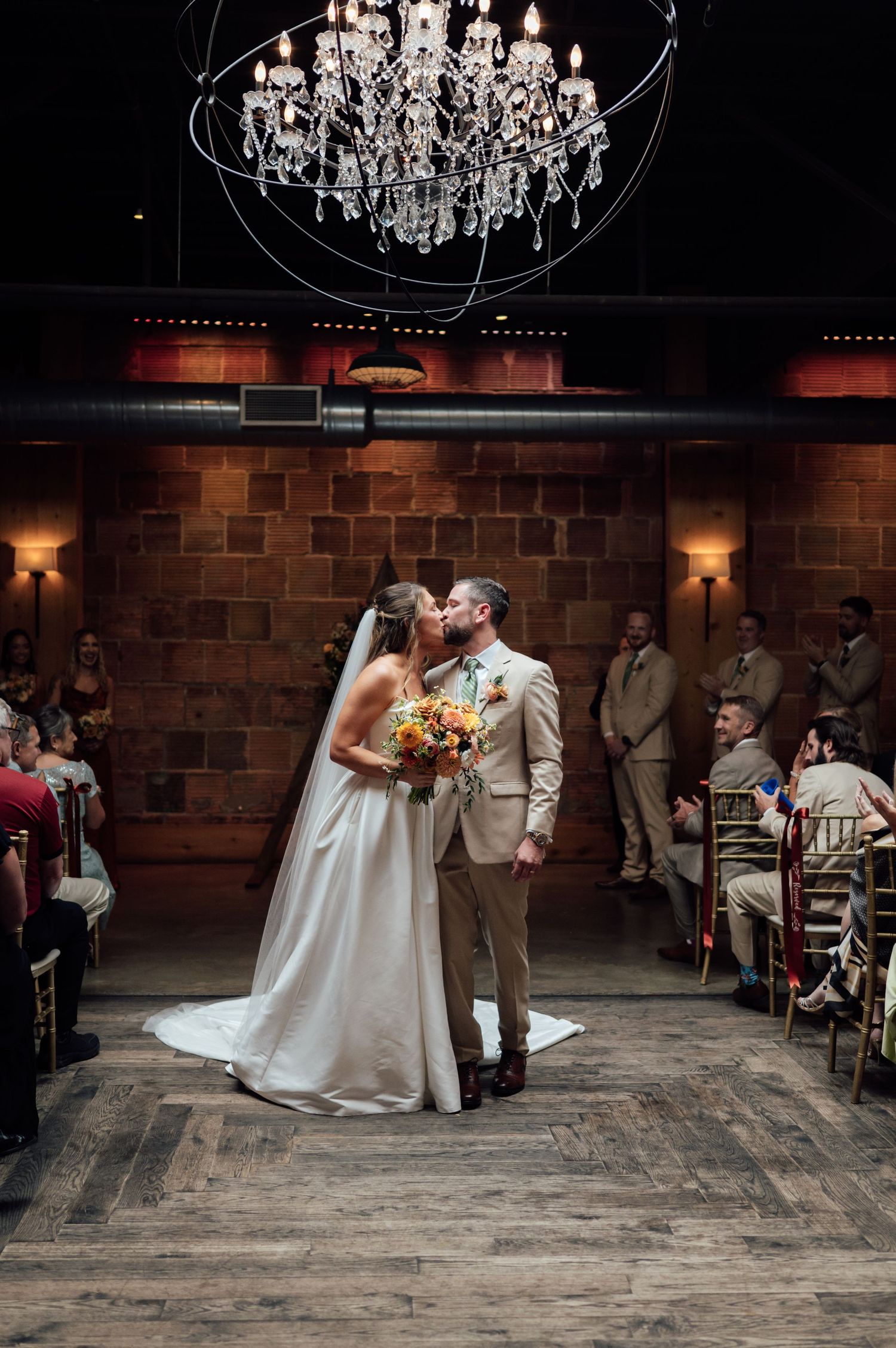 Bride and groom share first kiss at indoor wedding ceremony under crystal chandelier with brick walls and seated guests.