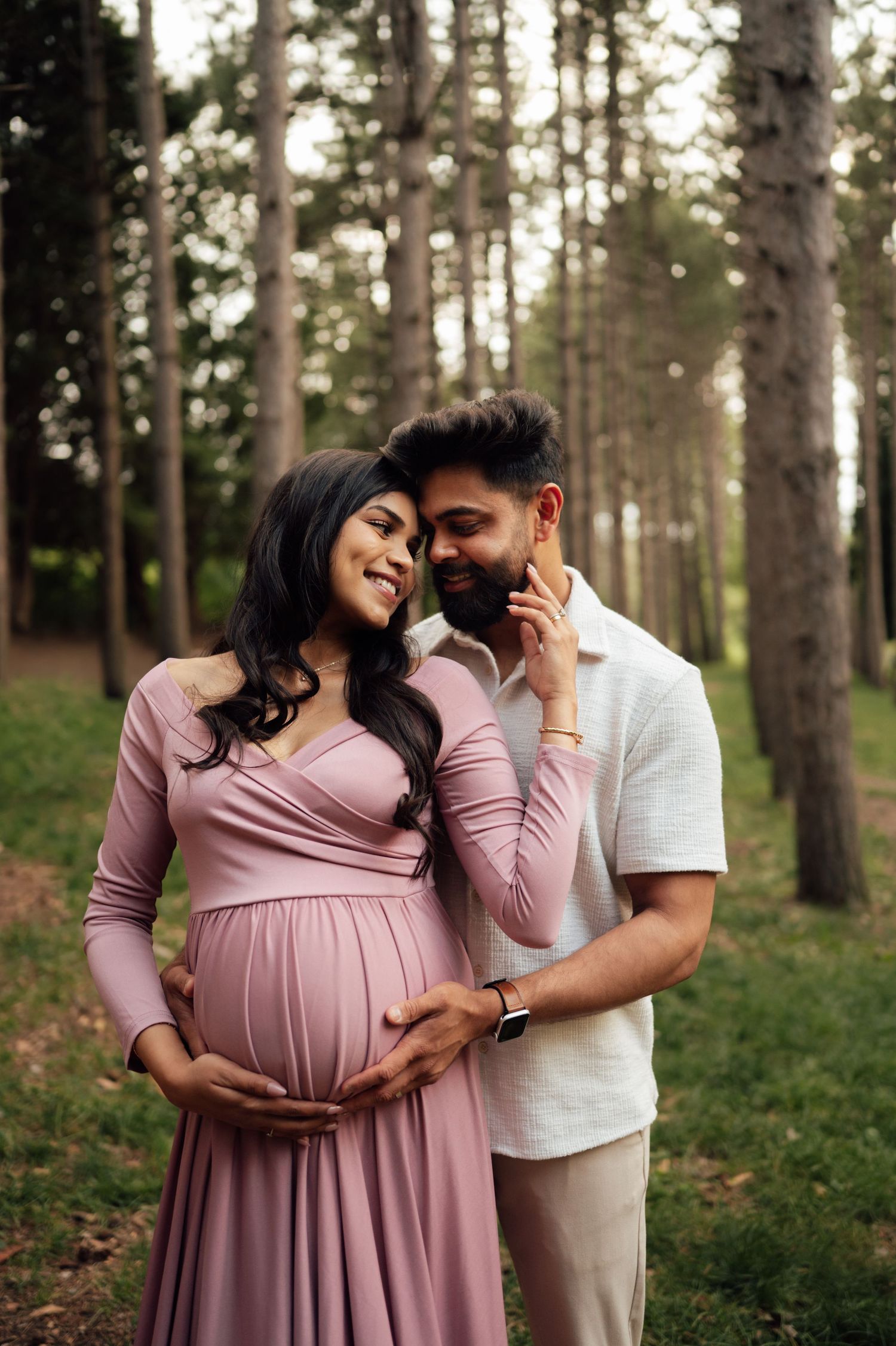 Pregnant couple embracing in a forest, woman in pink maxi dress cradling her bump while partner kisses her cheek.