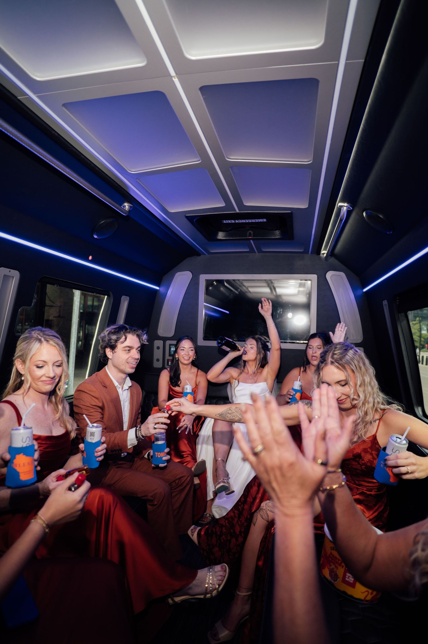 Wedding party celebrating in a party bus with blue LED lights, bridesmaids in red dresses raising drinks.