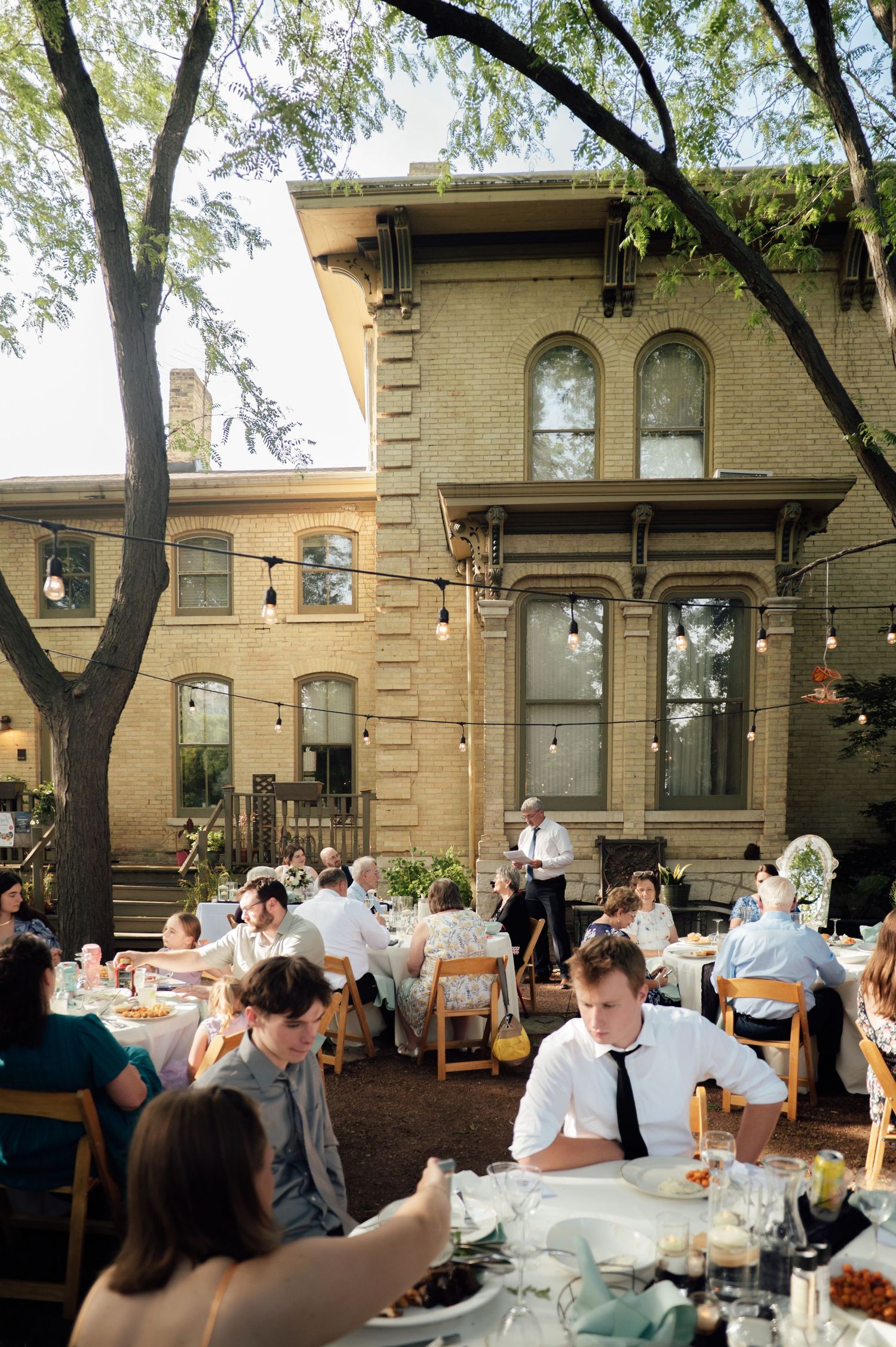 Outdoor wedding reception in a historic brick building courtyard with string lights and guests dining at round tables.
