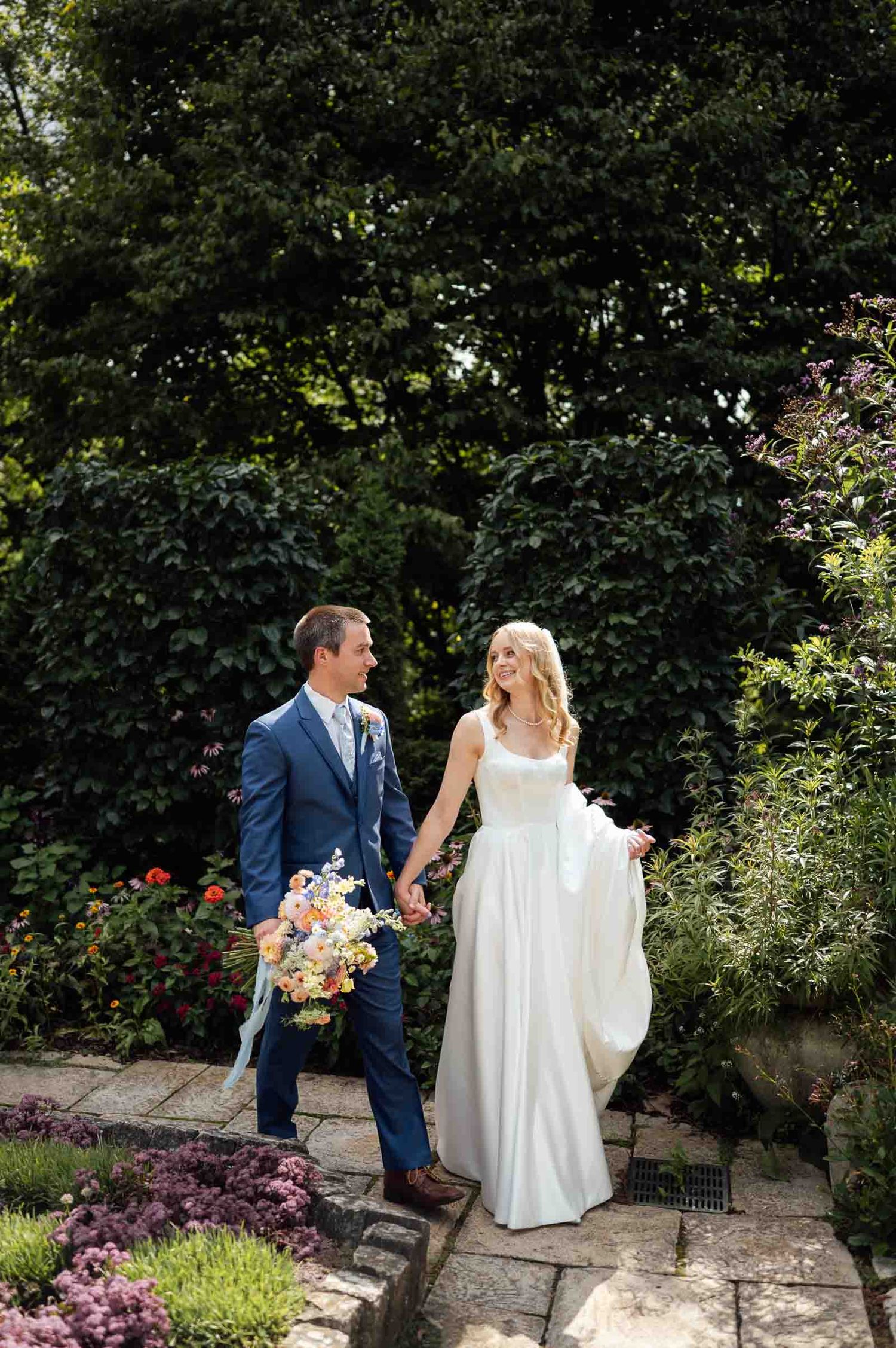 Bride in white gown and groom in blue suit walk hand-in-hand through lush garden holding colorful bouquet.