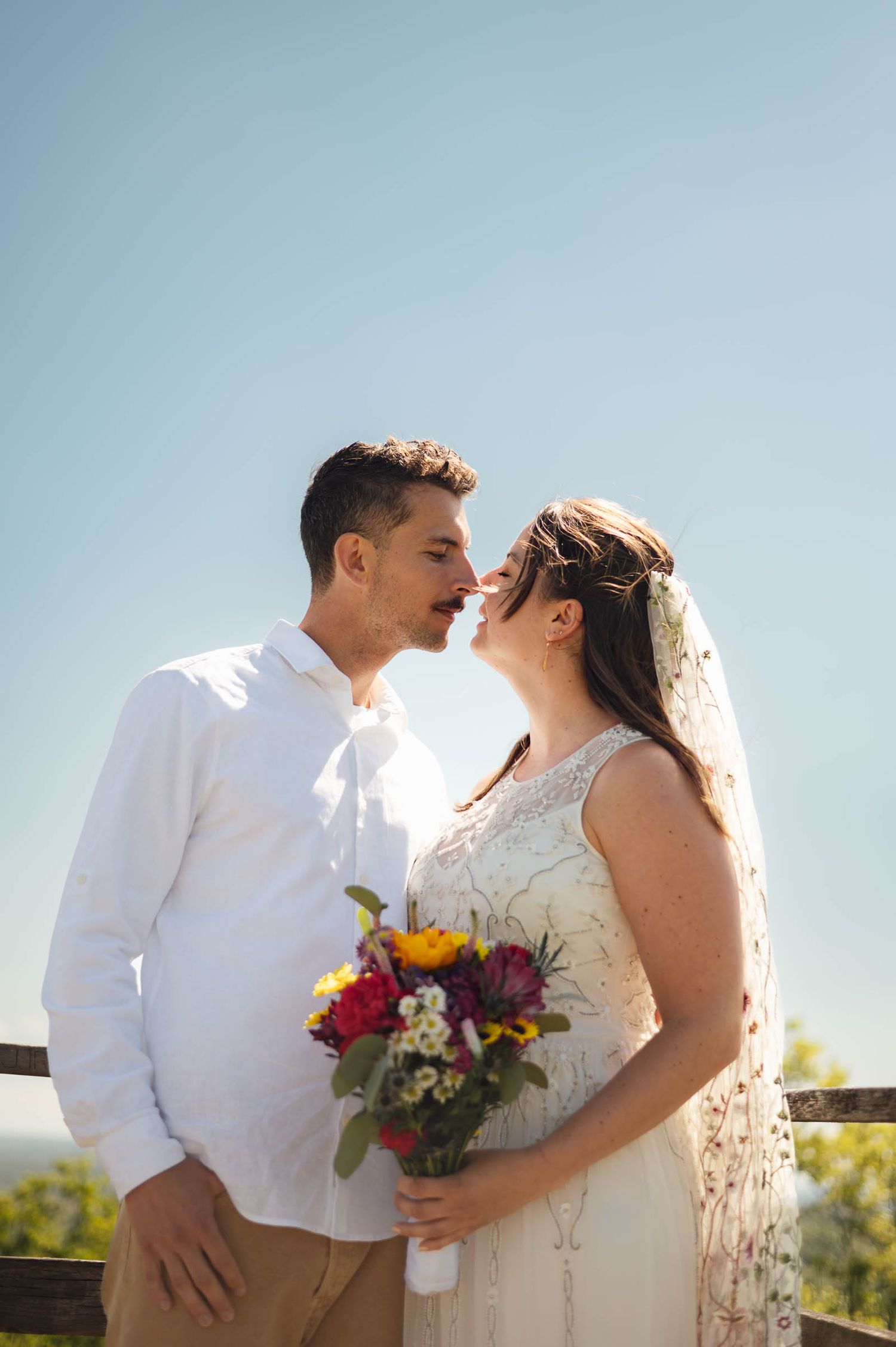 Bride and groom nearly kissing outdoors holding colorful bouquet under bright blue sky.