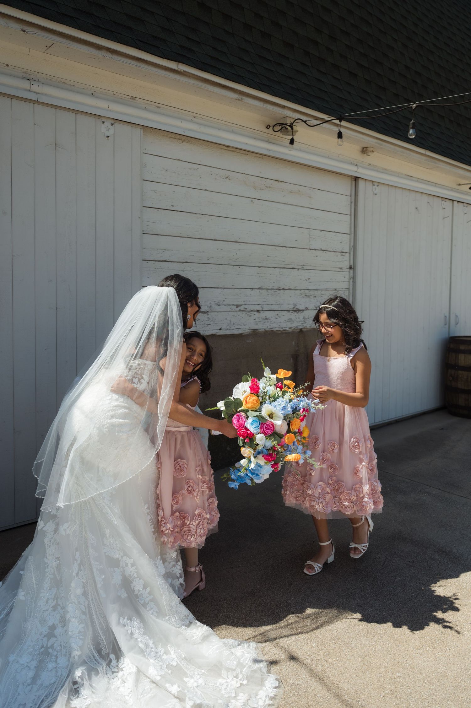 Someone in a white wedding dress bends down to interact with young attendants in pink dresses holding colorful flowers.