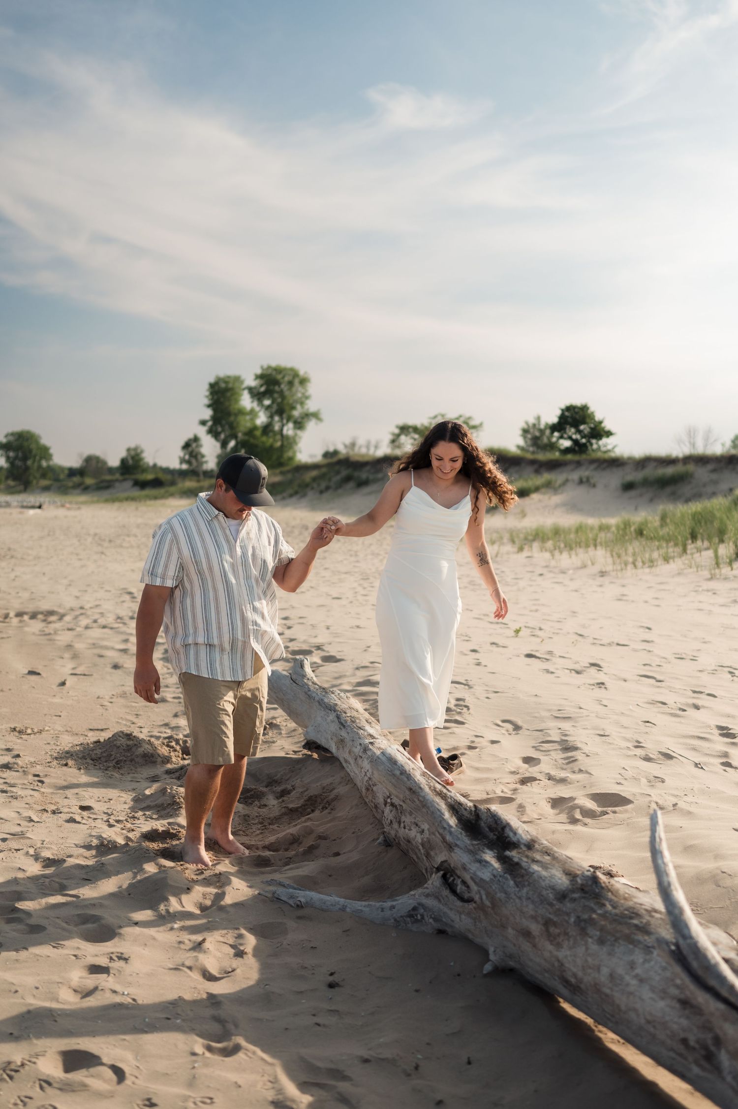 People walking and playing along a large piece of driftwood on a sandy beach during sunset.