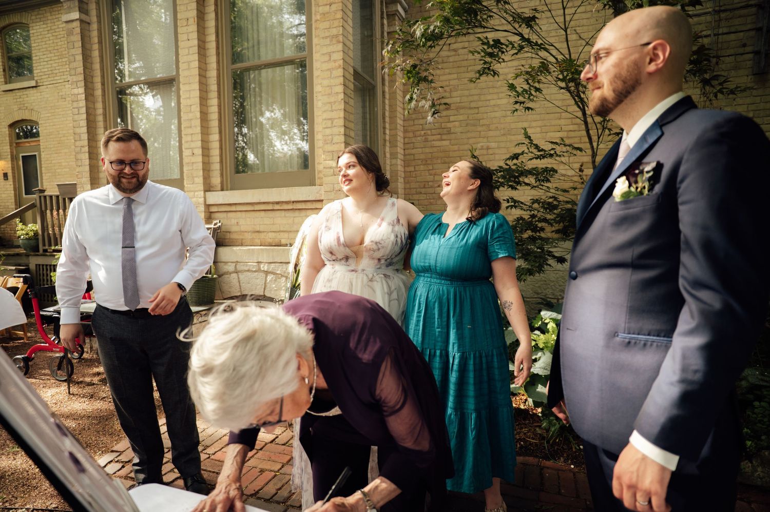 Wedding guests gather outdoors as someone signs a document during a formal ceremony near a brick building.