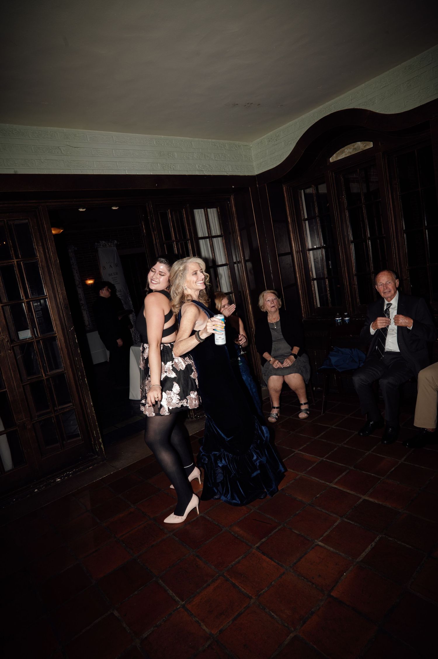 Two women in formal dresses dance together at wedding reception while guests watch nearby.