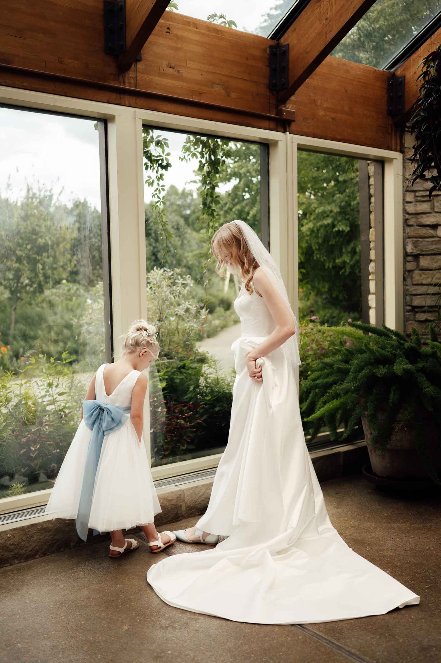 Bride in white gown with train stands with flower girl in blue bow dress by windows.