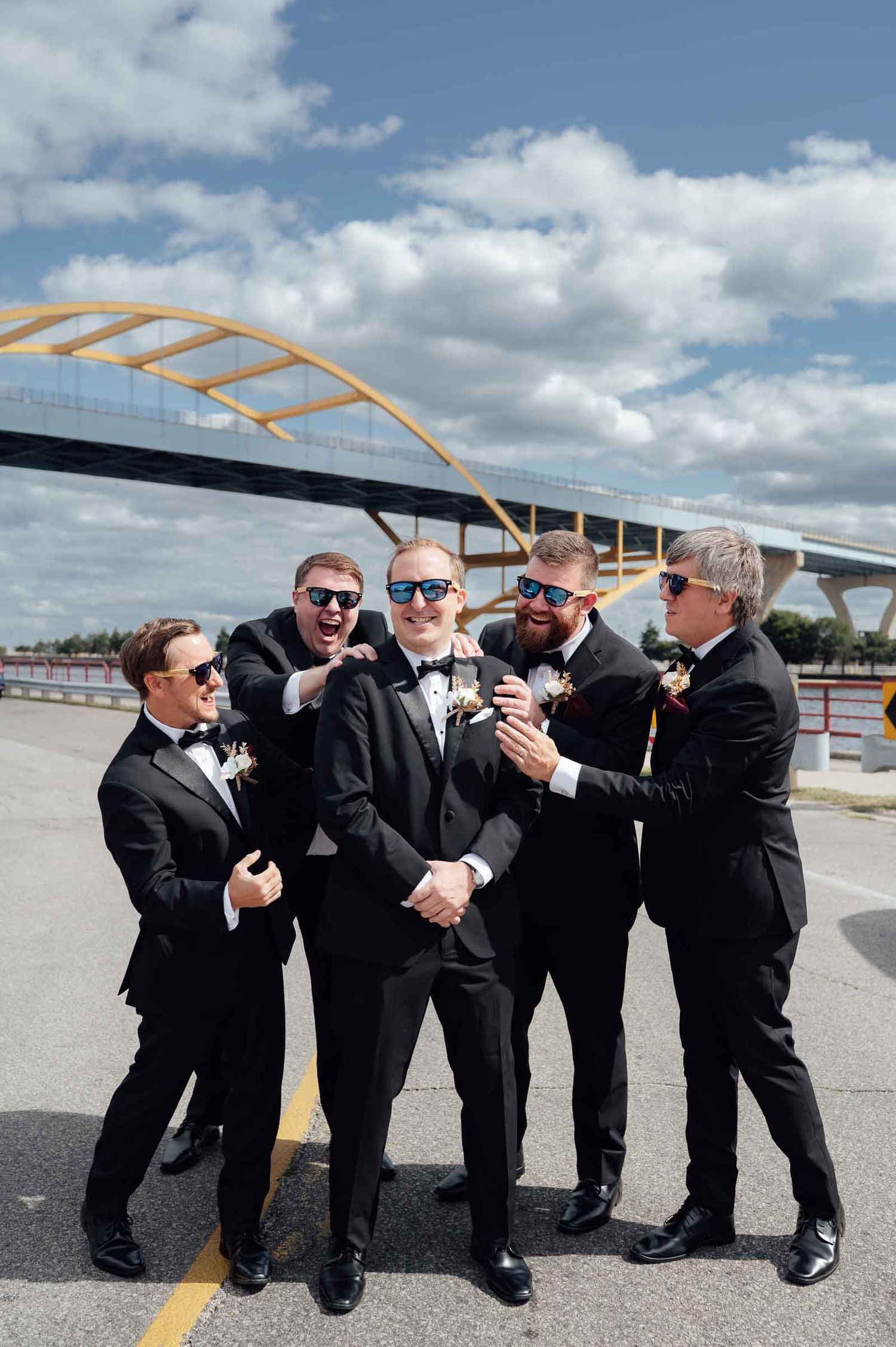 Groom and groomsmen in black tuxedos wearing sunglasses posing playfully near a yellow arch bridge outdoors.