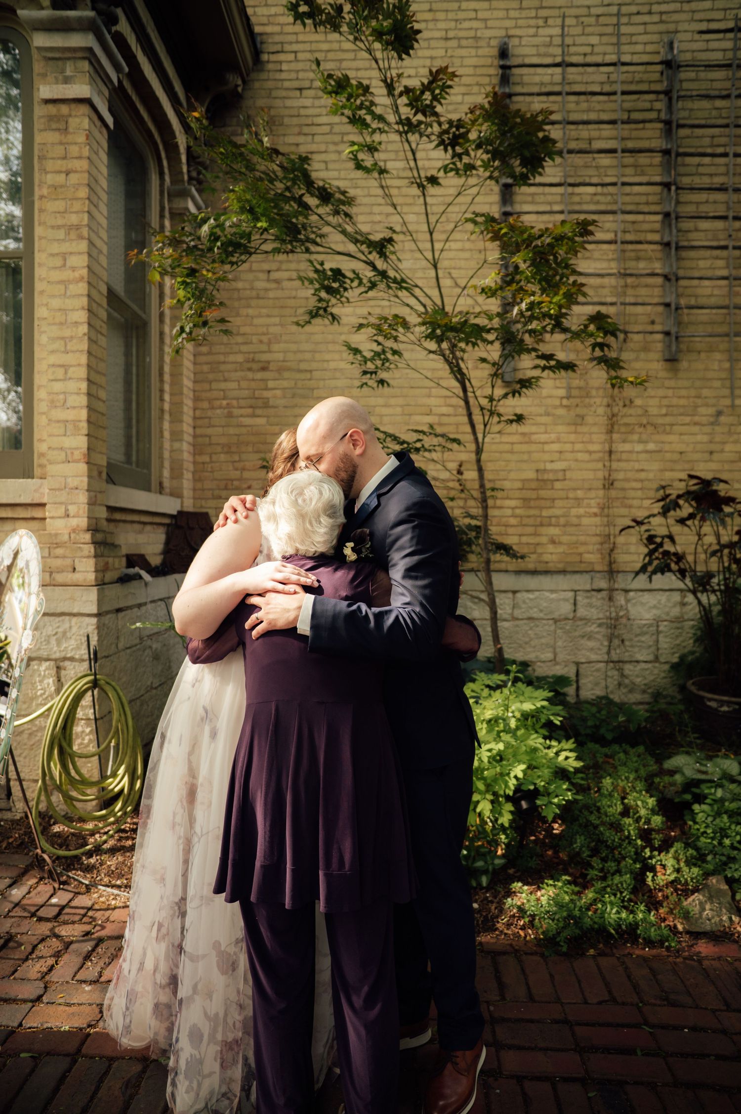 A bride and groom share a tender group hug with an elderly woman outside a brick building on their wedding day.