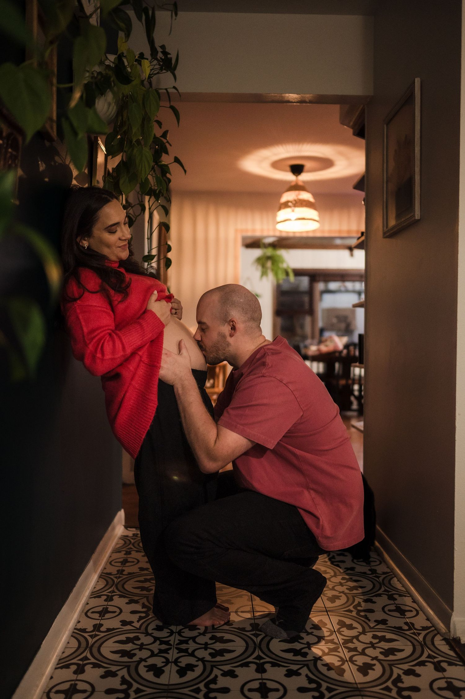 Man kneeling to kiss pregnant partner's baby bump in cozy home hallway with patterned tile floor.