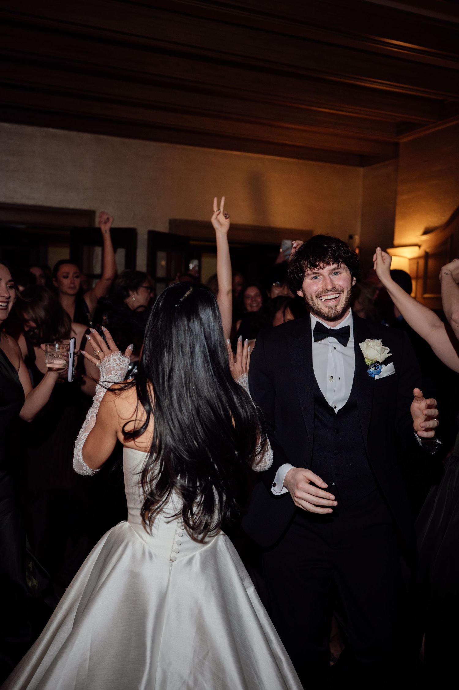 Smiling groom in tuxedo dances with bride in white ballgown surrounded by celebrating guests.