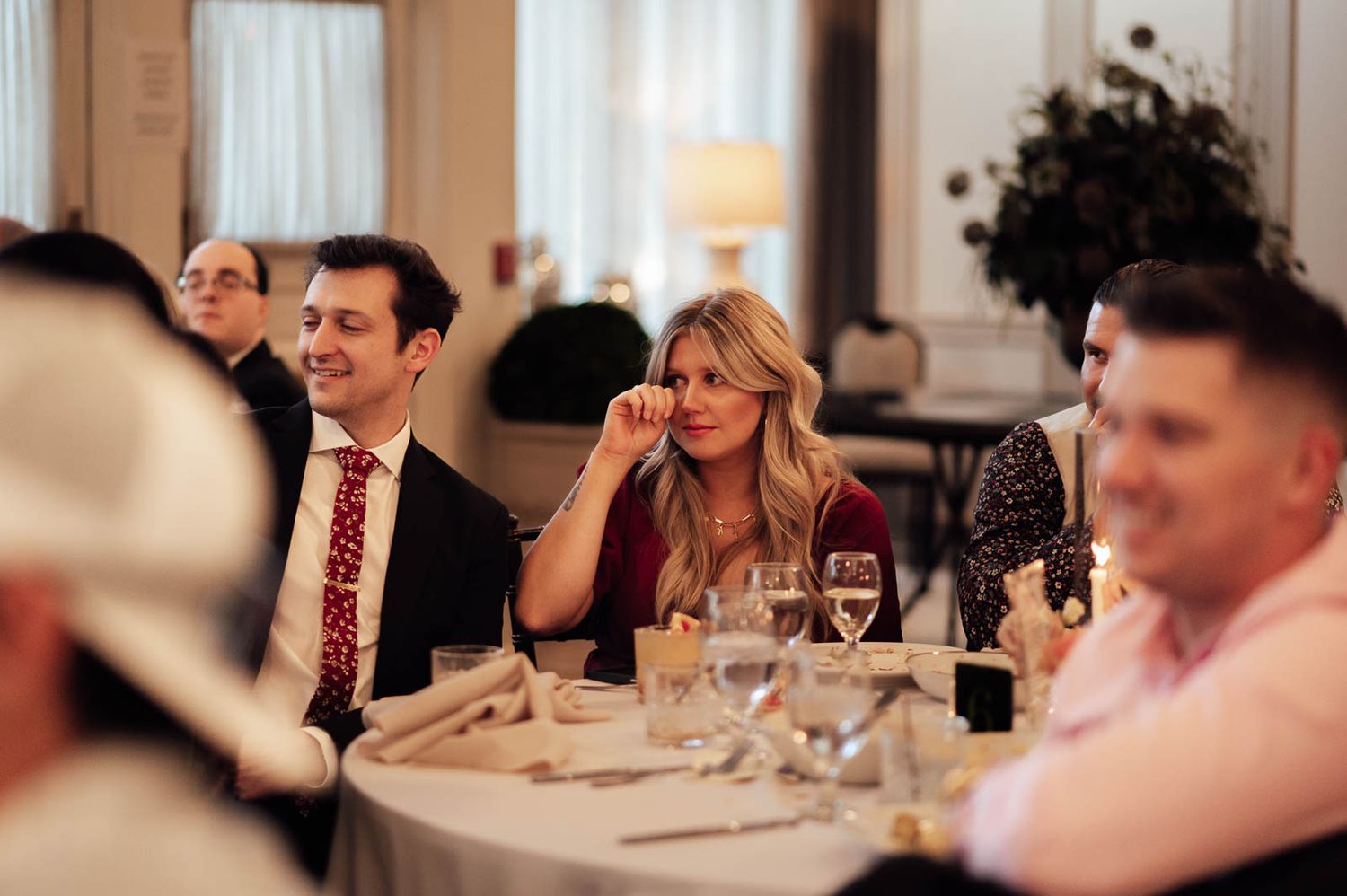 Wedding reception guests smiling and laughing at elegantly set tables with white linens and glassware.
