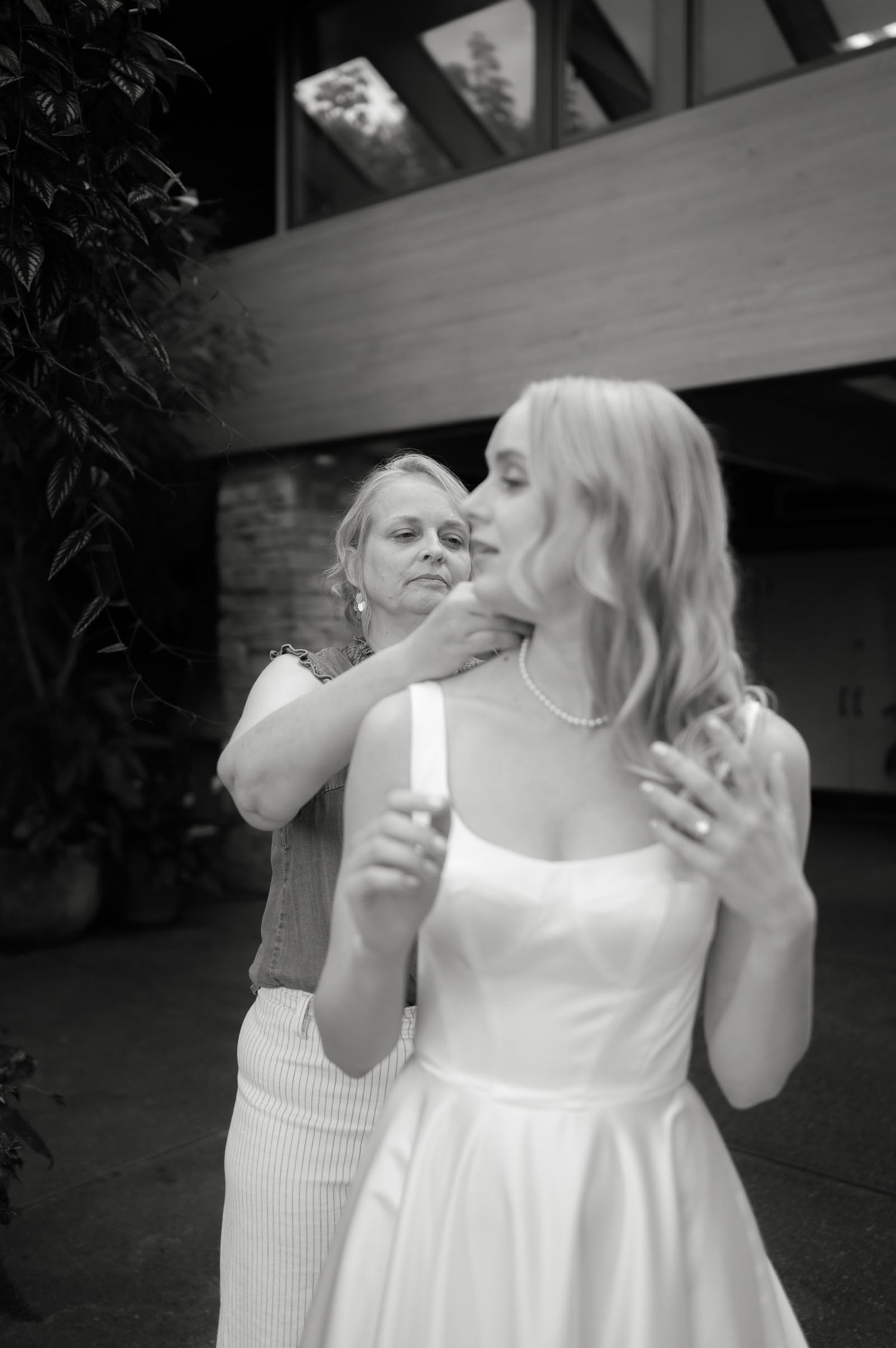 Black and white photo of mother fastening necklace on bride before wedding ceremony.