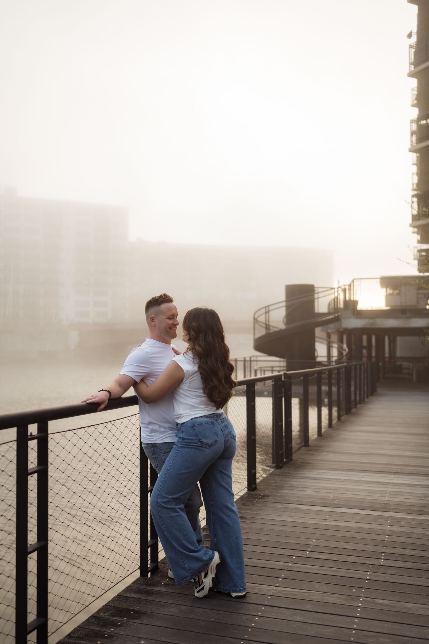 Couple in matching white tees and jeans embrace on a foggy waterfront boardwalk at sunrise.