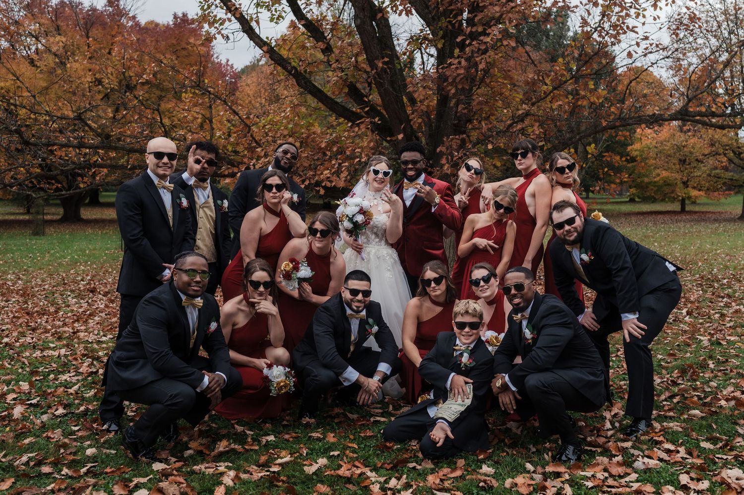 A group of wedding party members in burgundy and black attire pose together on autumn leaves in a park setting.