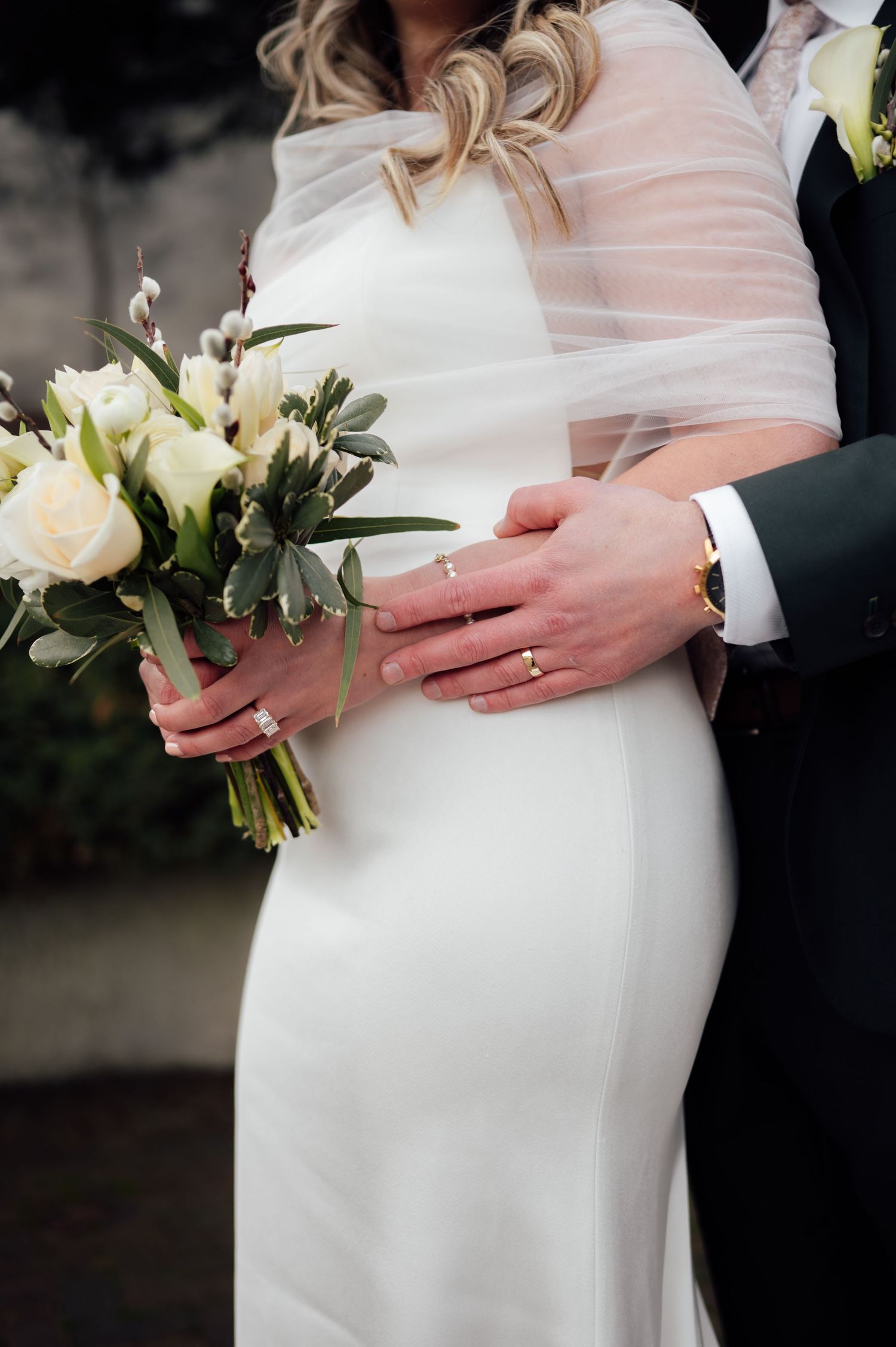 Close-up of bride and groom hands with wedding rings, bride holding white roses and greenery bouquet.