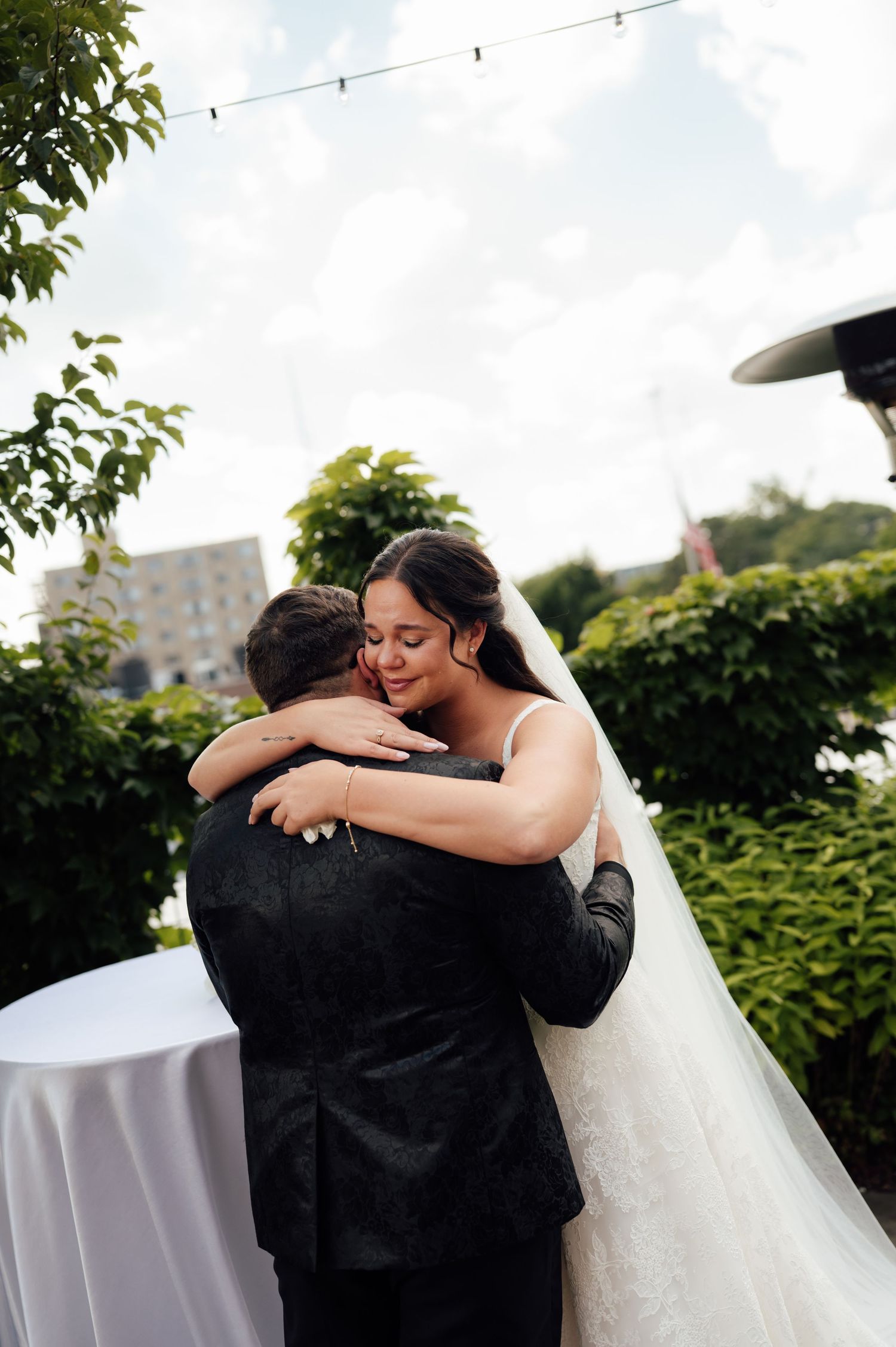 Bride in white gown embracing groom in black suit outdoors at wedding reception with lush greenery backdrop.