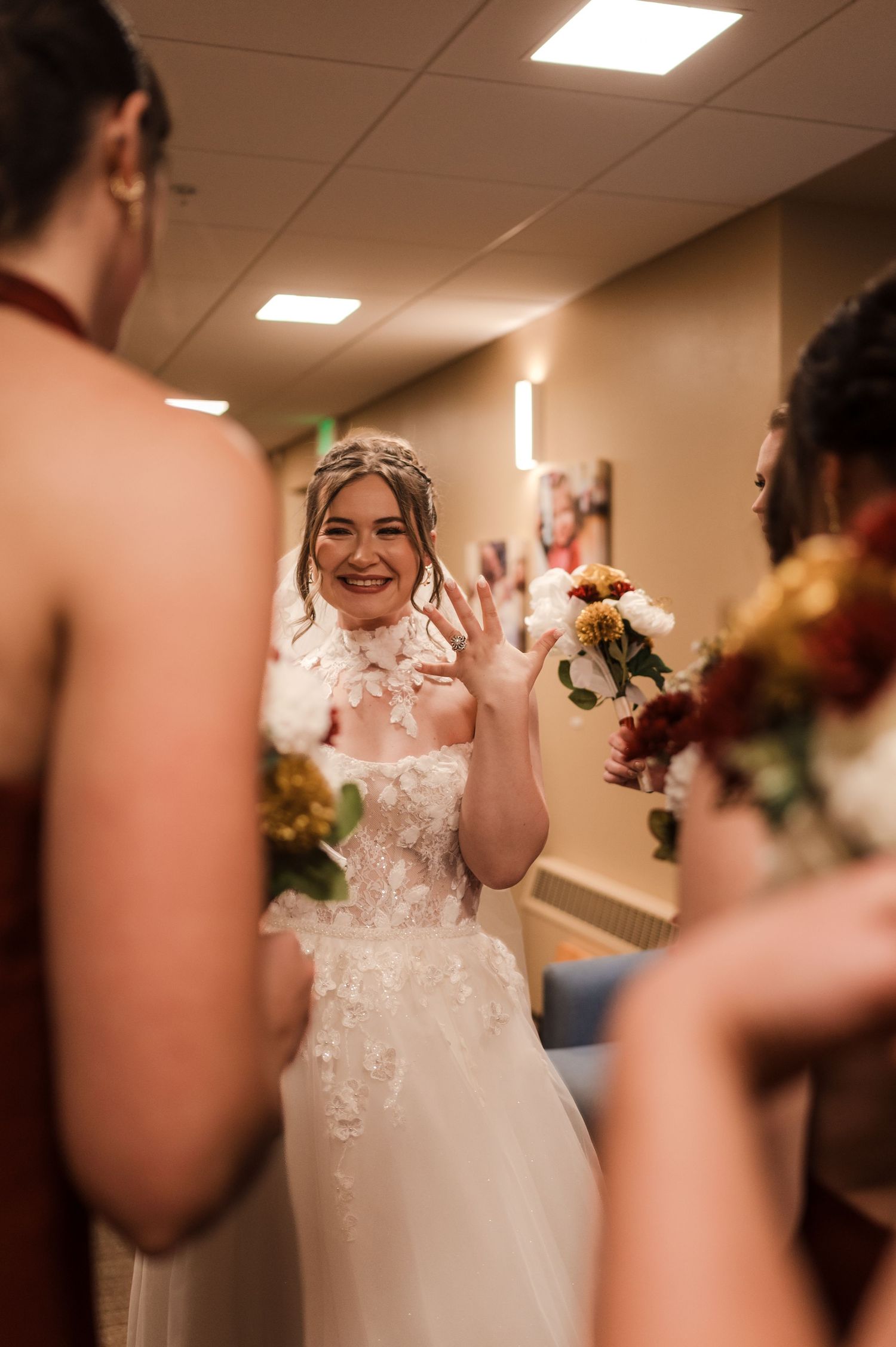 A bride in a white lace wedding dress shows off her ring during an emotional first look moment in a church hallway.