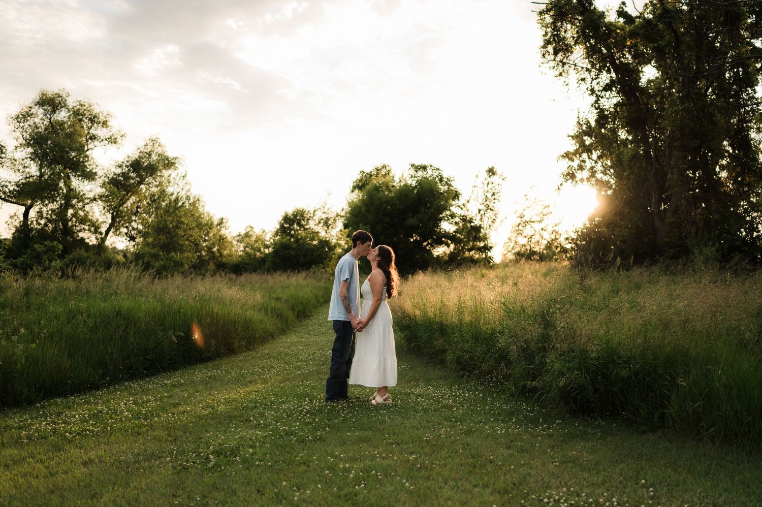 Couple sharing a tender kiss on a lush green path surrounded by trees at golden hour sunset.