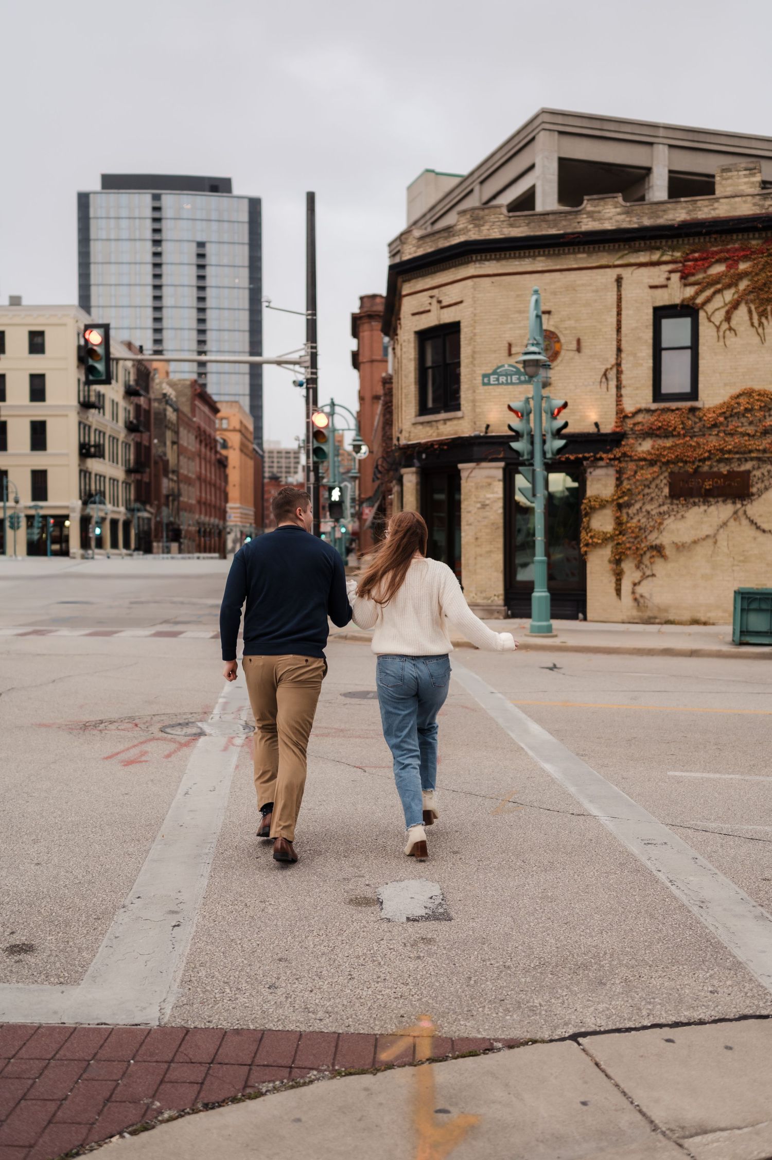Couple walking hand-in-hand across a city street intersection with historic brick buildings in fall.