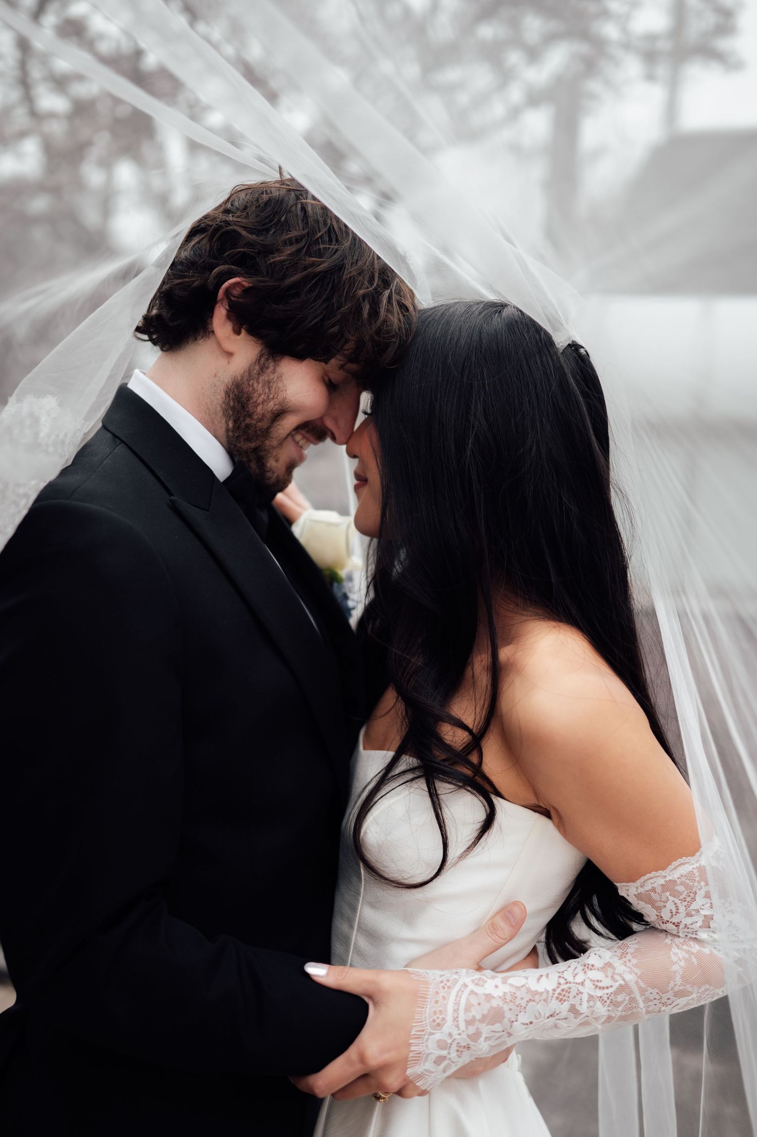 Romantic bride and groom share intimate moment under flowing veil, foreheads touching tenderly.