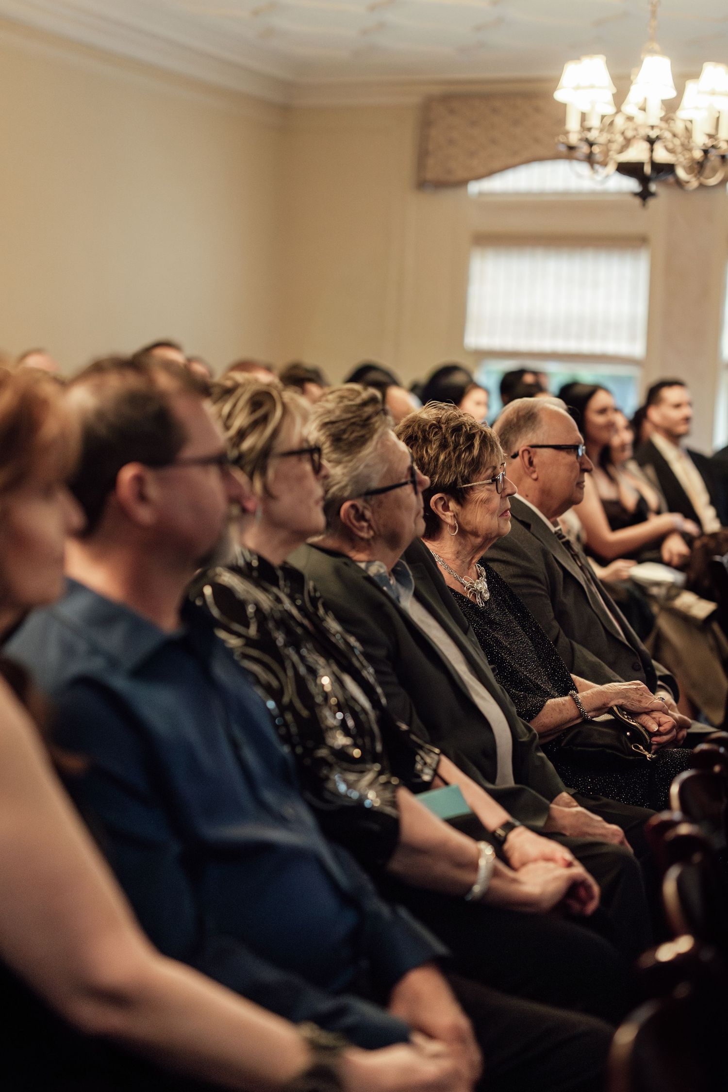 Wedding guests seated in elegant room with chandelier, smiling and watching ceremony unfold.