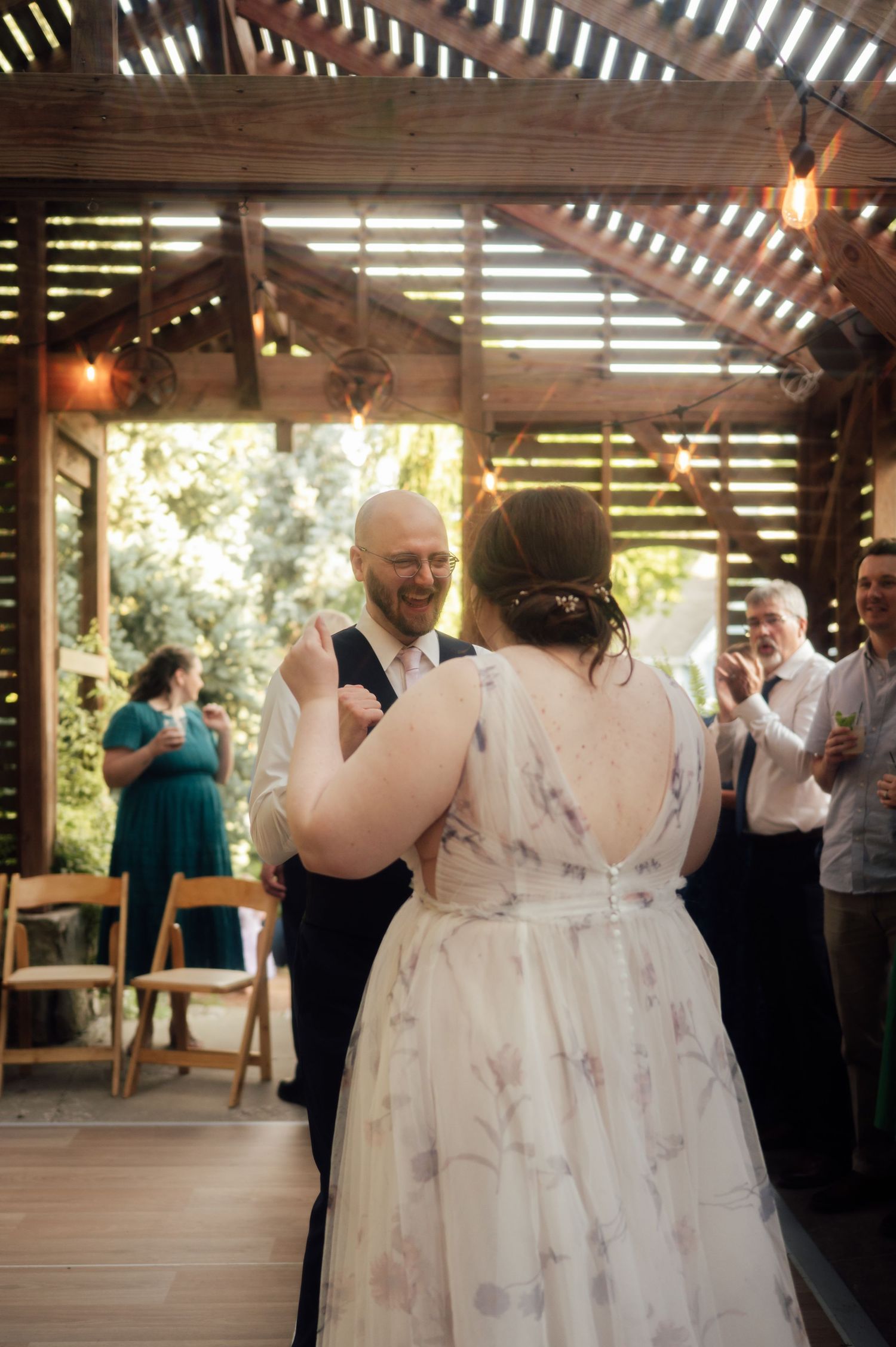 Bride and groom share first dance under rustic wooden pergola with string lights, guests applauding at outdoor wedding reception.