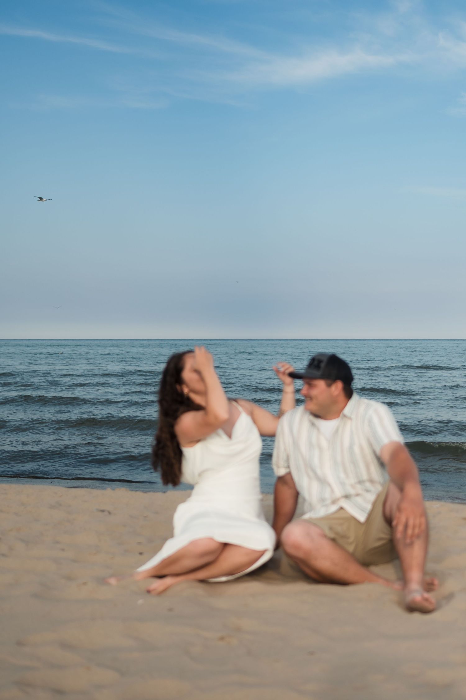 Couple sitting together on a sandy beach laughing with the ocean waves behind them.
