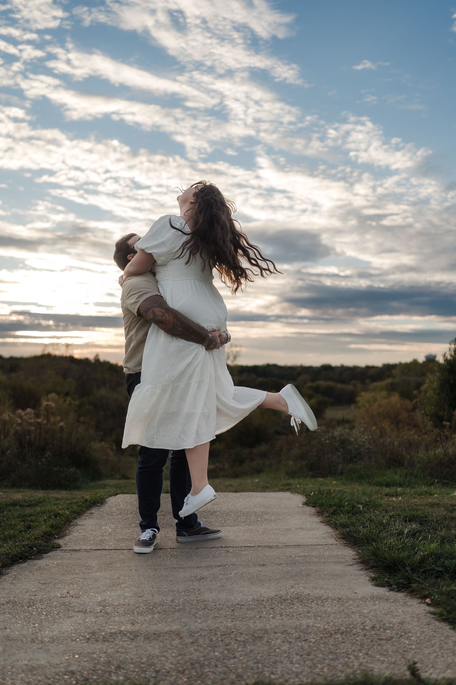 Couple sharing a romantic embrace on a path at sunset, woman lifted off ground with hair flowing in the breeze.