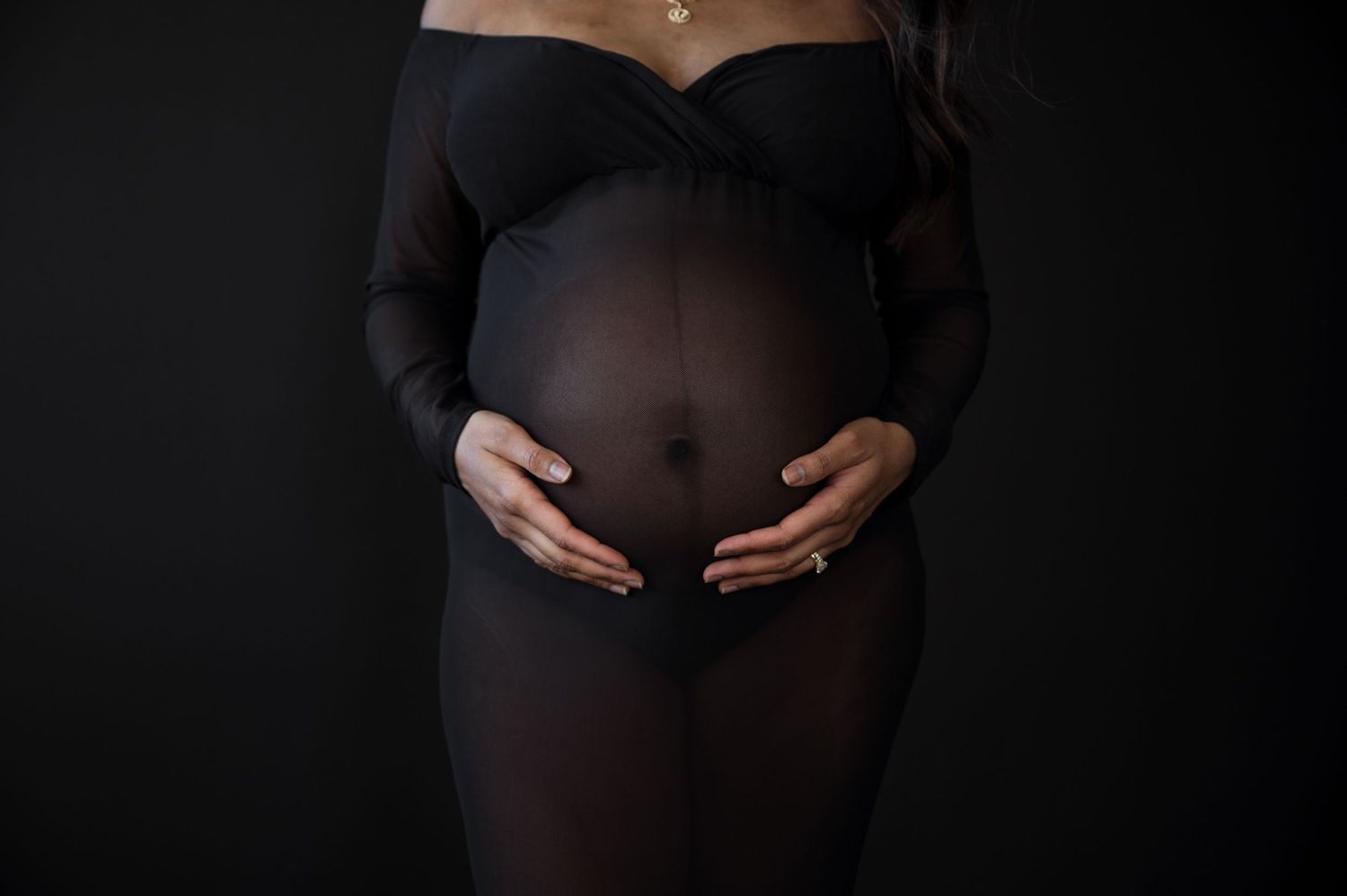 Close-up of pregnant belly in sheer black bodysuit with hands cradling bump.