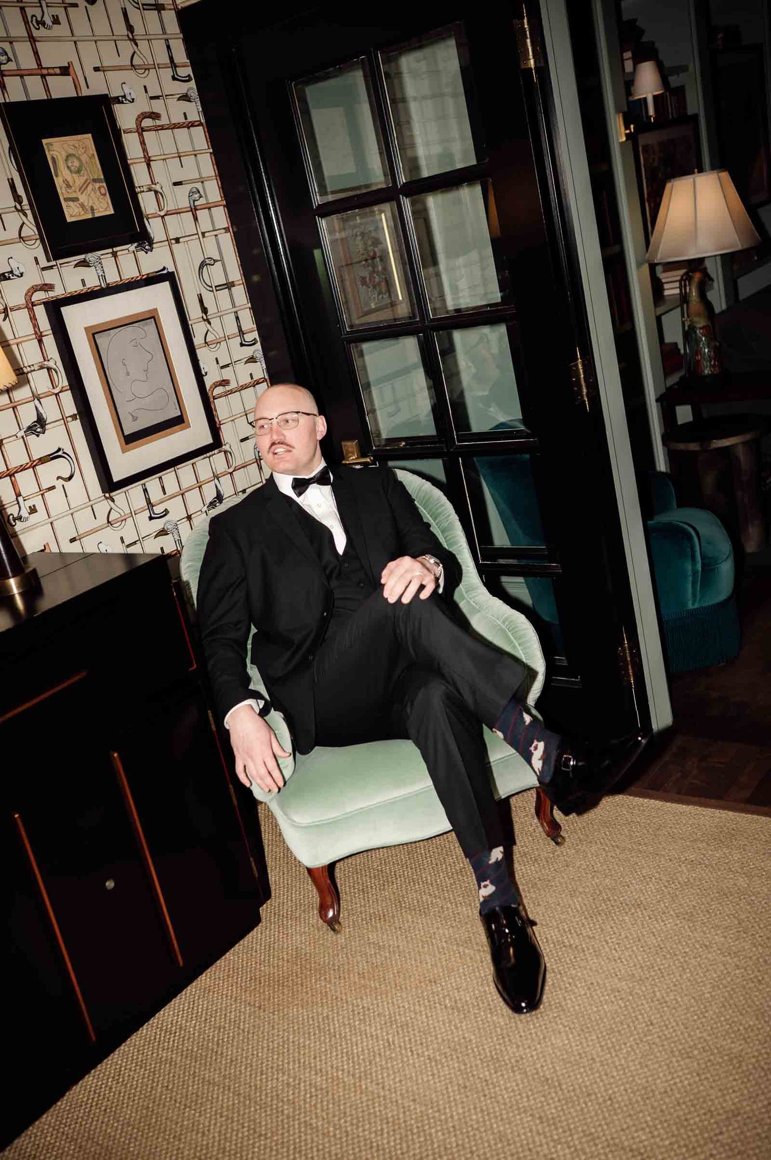 Elegant bald man in black tuxedo and bow tie sitting in mint green chair in stylish hotel lounge.