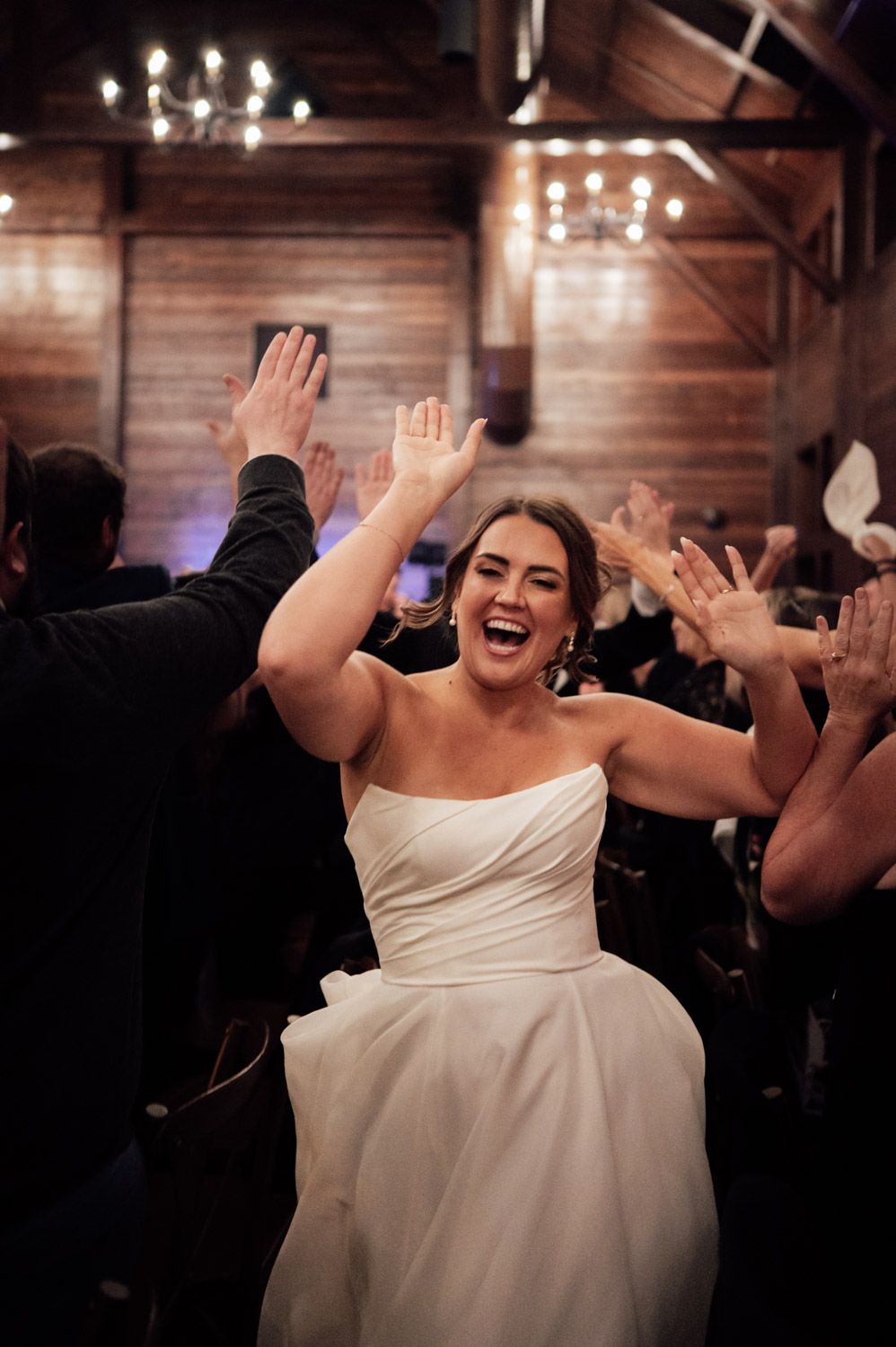 Joyful bride in white strapless gown celebrating with raised hands during wedding reception entrance.