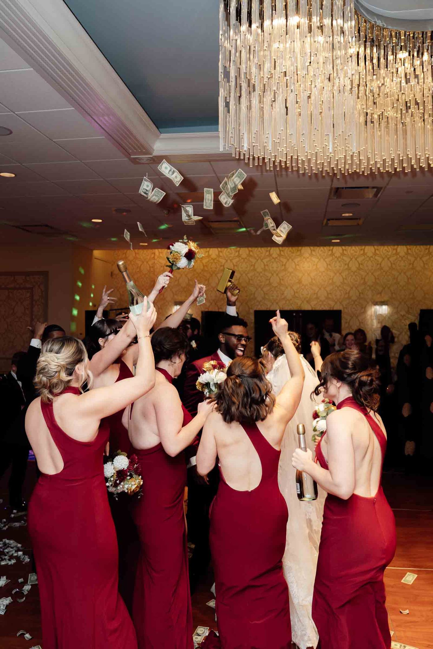 Wedding party in red dresses tosses money in the air under a crystal chandelier at reception.