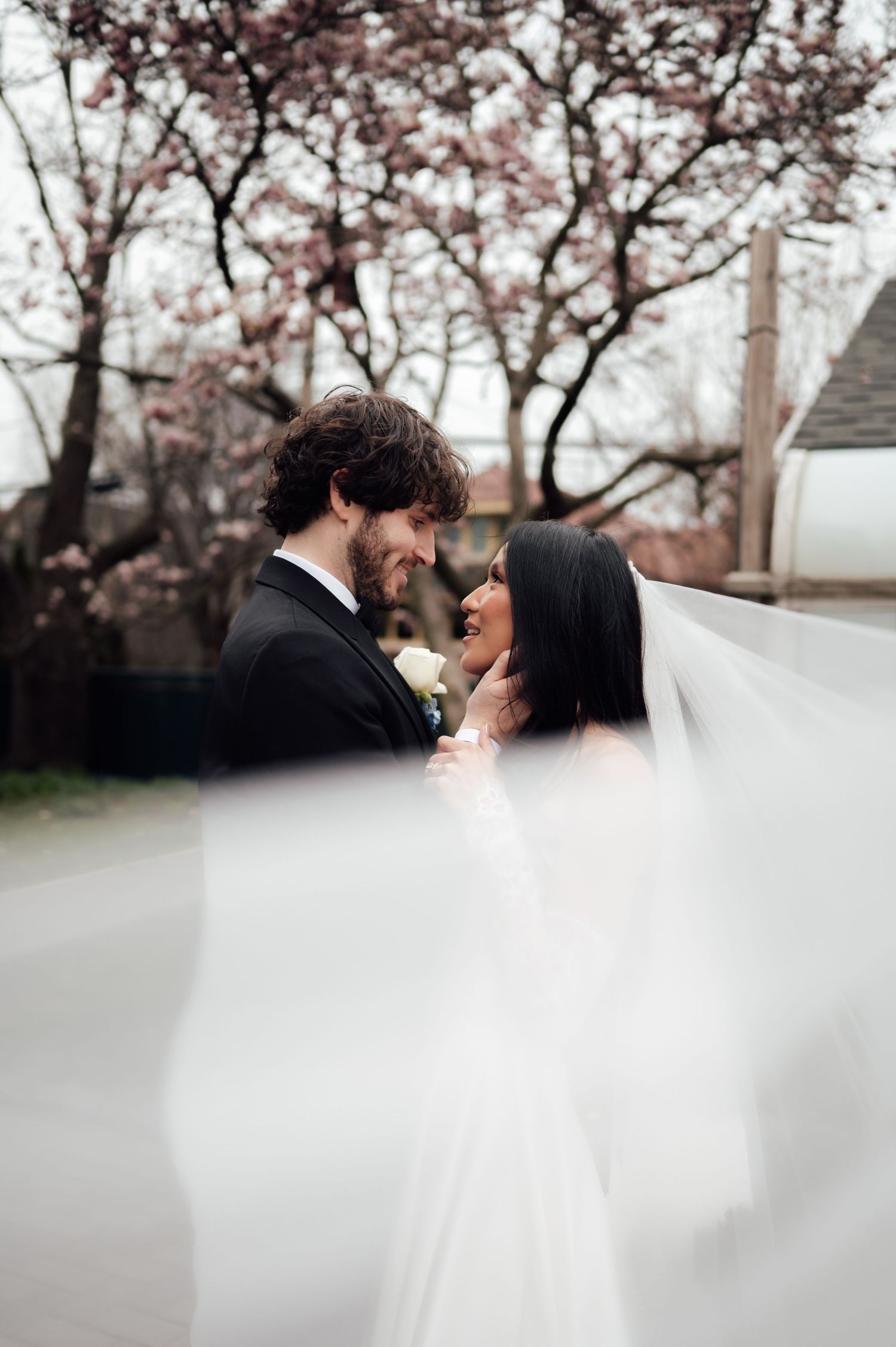 Bride and groom share an intimate moment under a flowing veil with blooming cherry trees in the background.