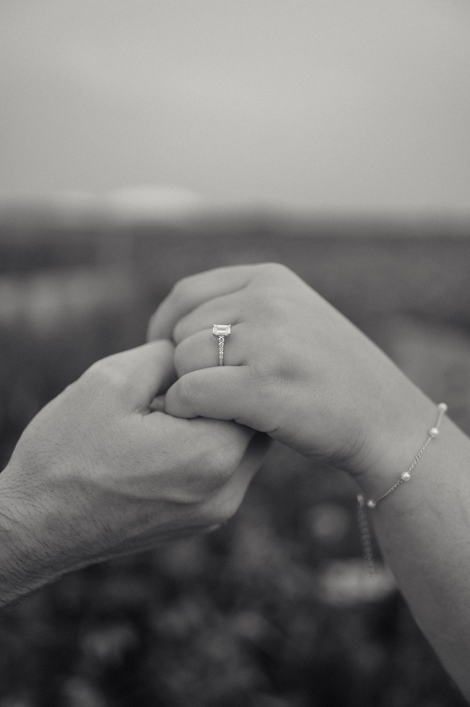 A close-up black and white photo of two hands clasped together, with an engagement ring visible on one hand.