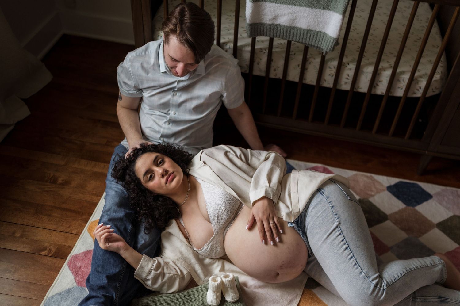 A pregnant woman lies on a patterned blanket on hardwood floor while someone sits nearby in a peaceful moment at home.