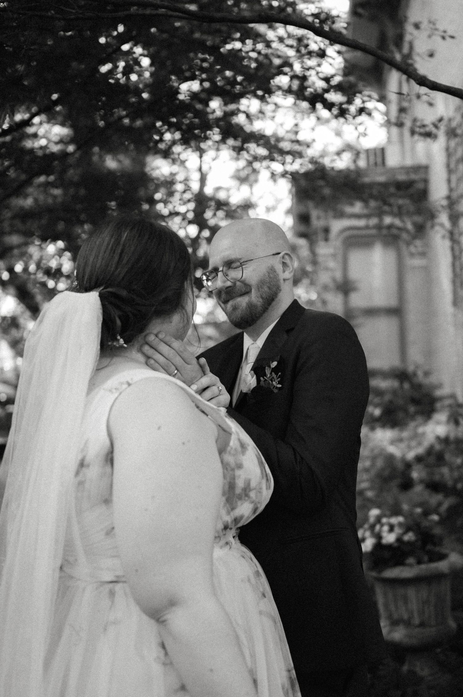 Black and white wedding photo of groom tenderly cupping brides face while smiling outdoors near historic building.