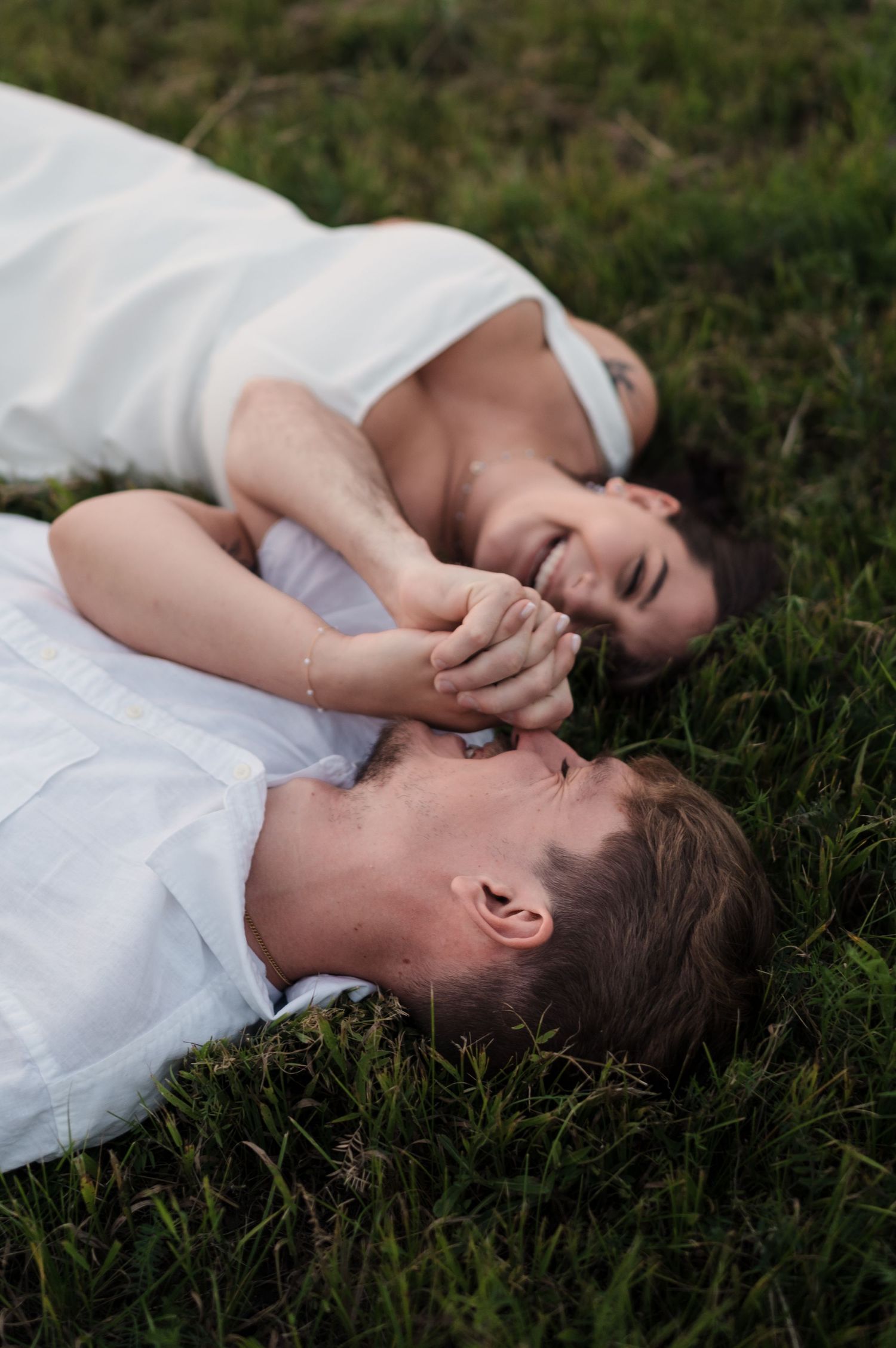 Couple dressed in white lying on green grass holding hands and laughing together.