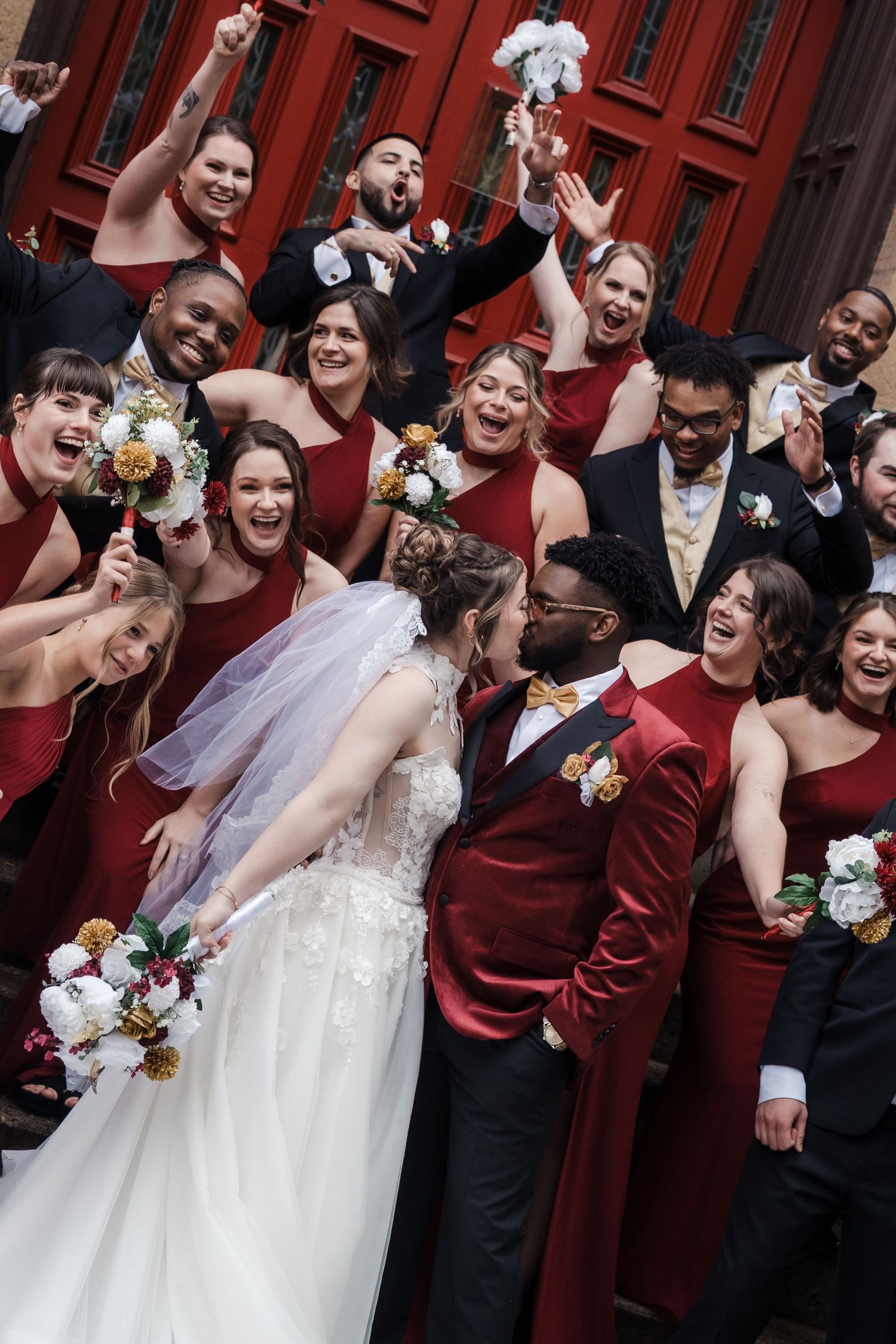 Wedding party in burgundy and black attire laughs joyfully against red doors at celebration.