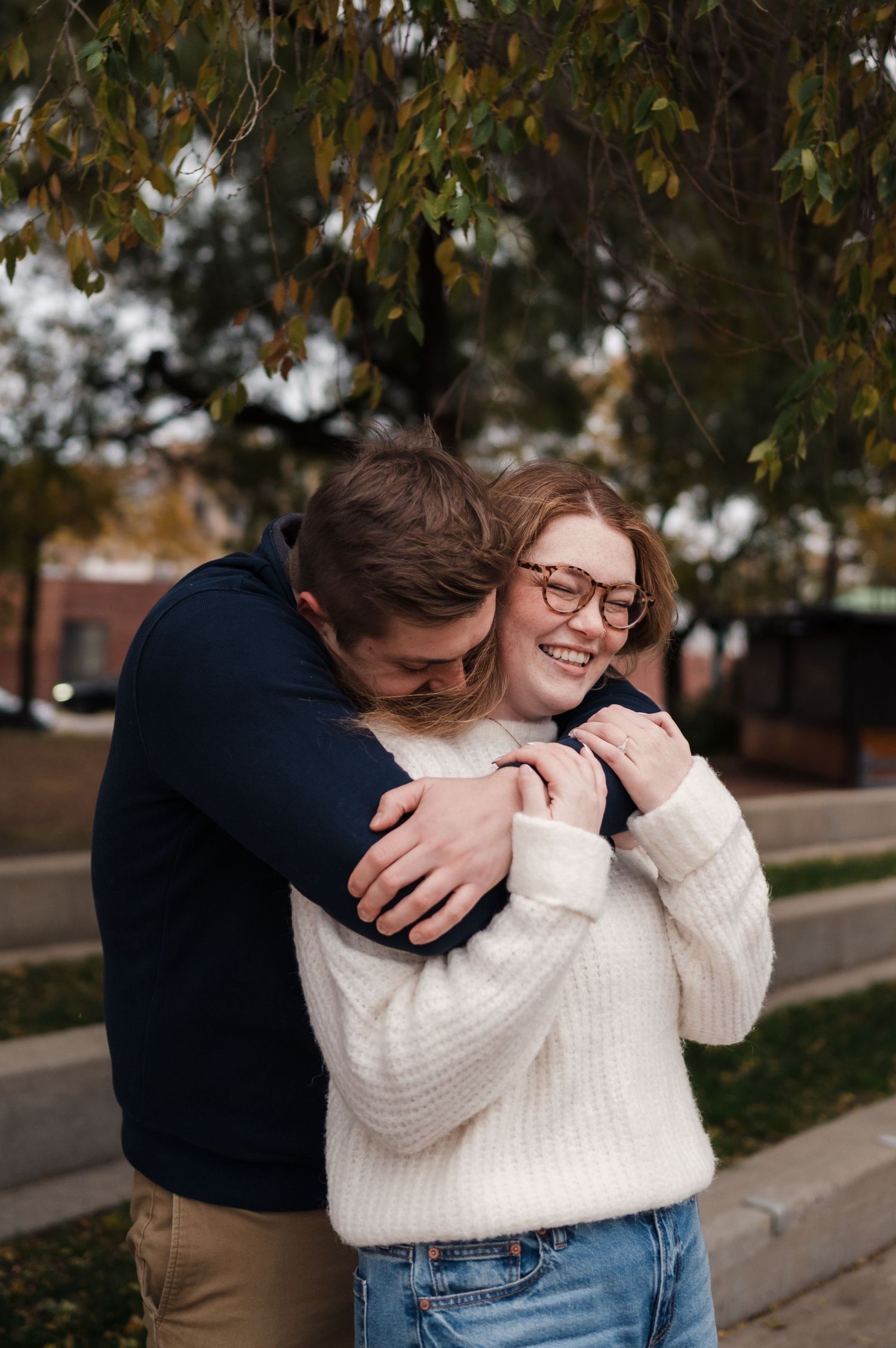 Man hugging woman from behind as she laughs joyfully under autumn trees in urban park.