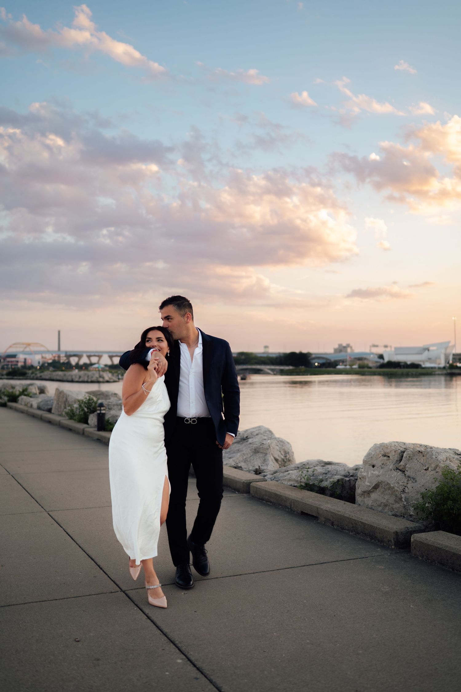 Couple walking along waterfront promenade at sunset, woman in white dress and man in navy suit, colorful sky.