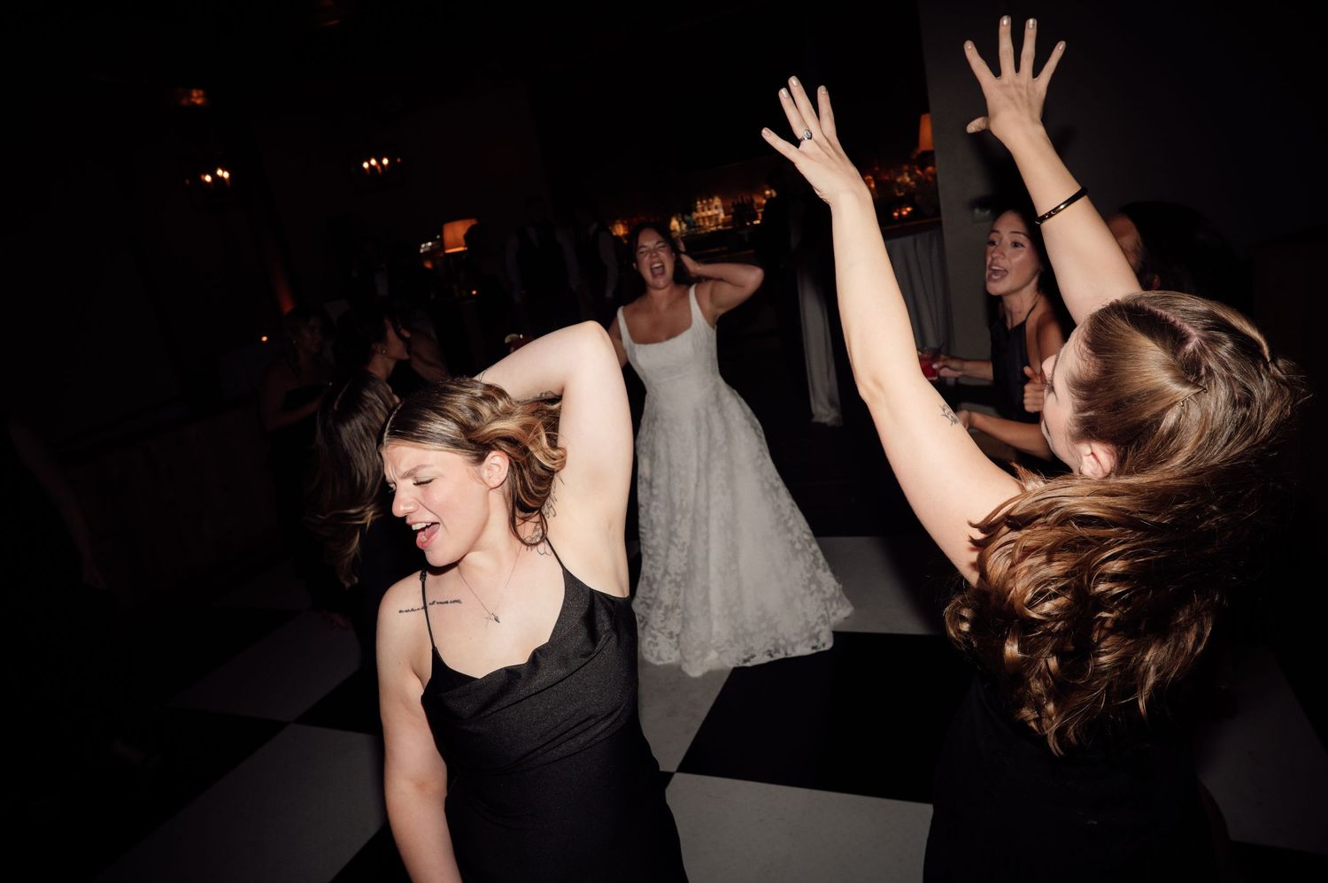 Women dancing joyfully at a wedding reception, with a bride in white gown visible in the background on a black and white dance floor.