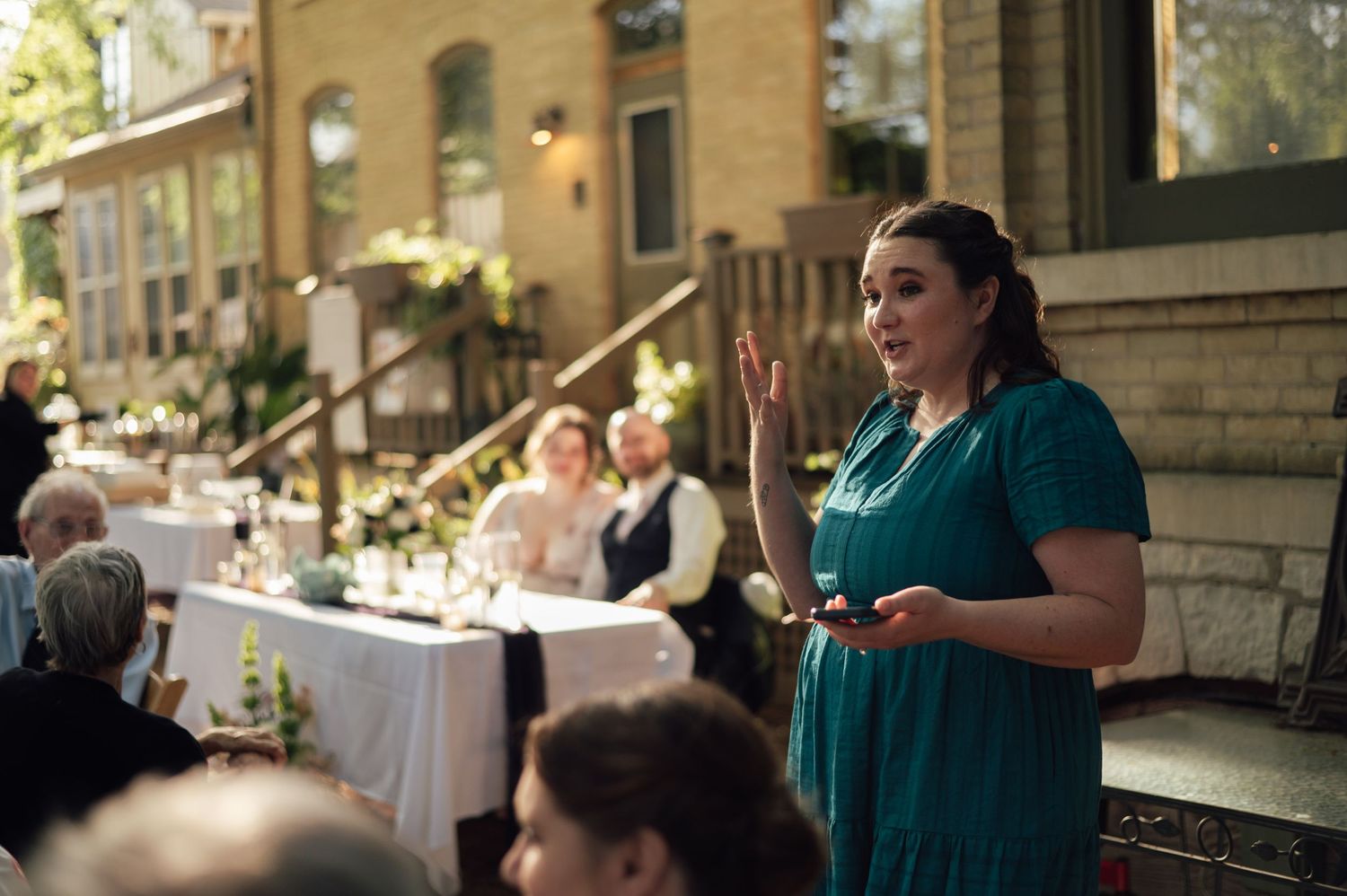 Woman in teal dress giving a speech at an elegant outdoor wedding reception with guests seated at decorated tables.