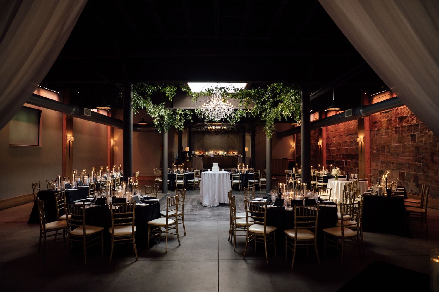 Elegant wedding reception venue with dark ambiance, chandelier, greenery ceiling, gold chairs, and black tablecloths.
