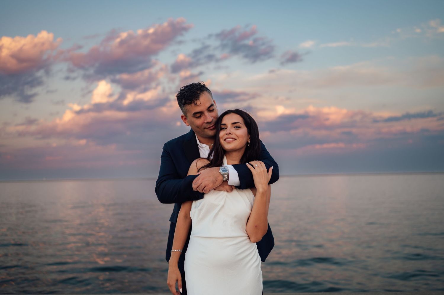 Romantic couple embracing at sunset by the ocean, groom in navy suit kissing bride in white wedding dress.