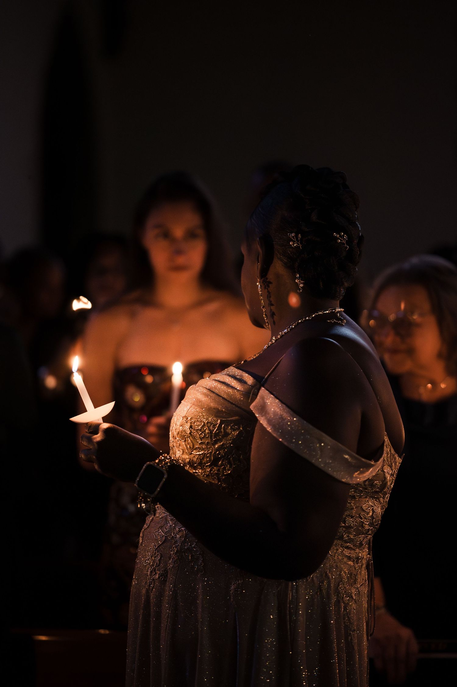 A candlelit ceremony shows silhouettes of people in formal attire holding glowing candles in a darkened room.