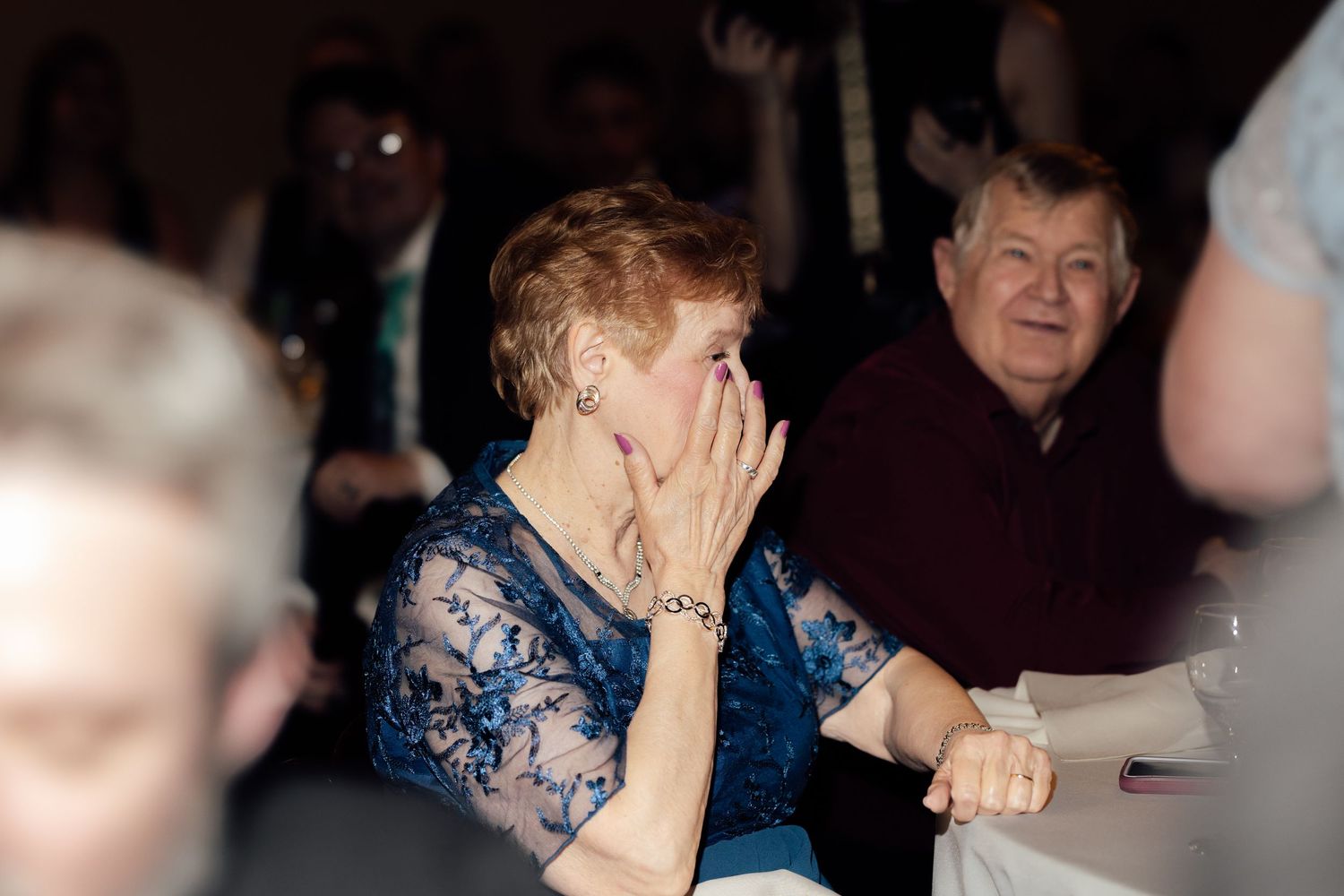 Elderly woman covering her mouth in surprise at a formal event, seated next to an older man in a dark venue.