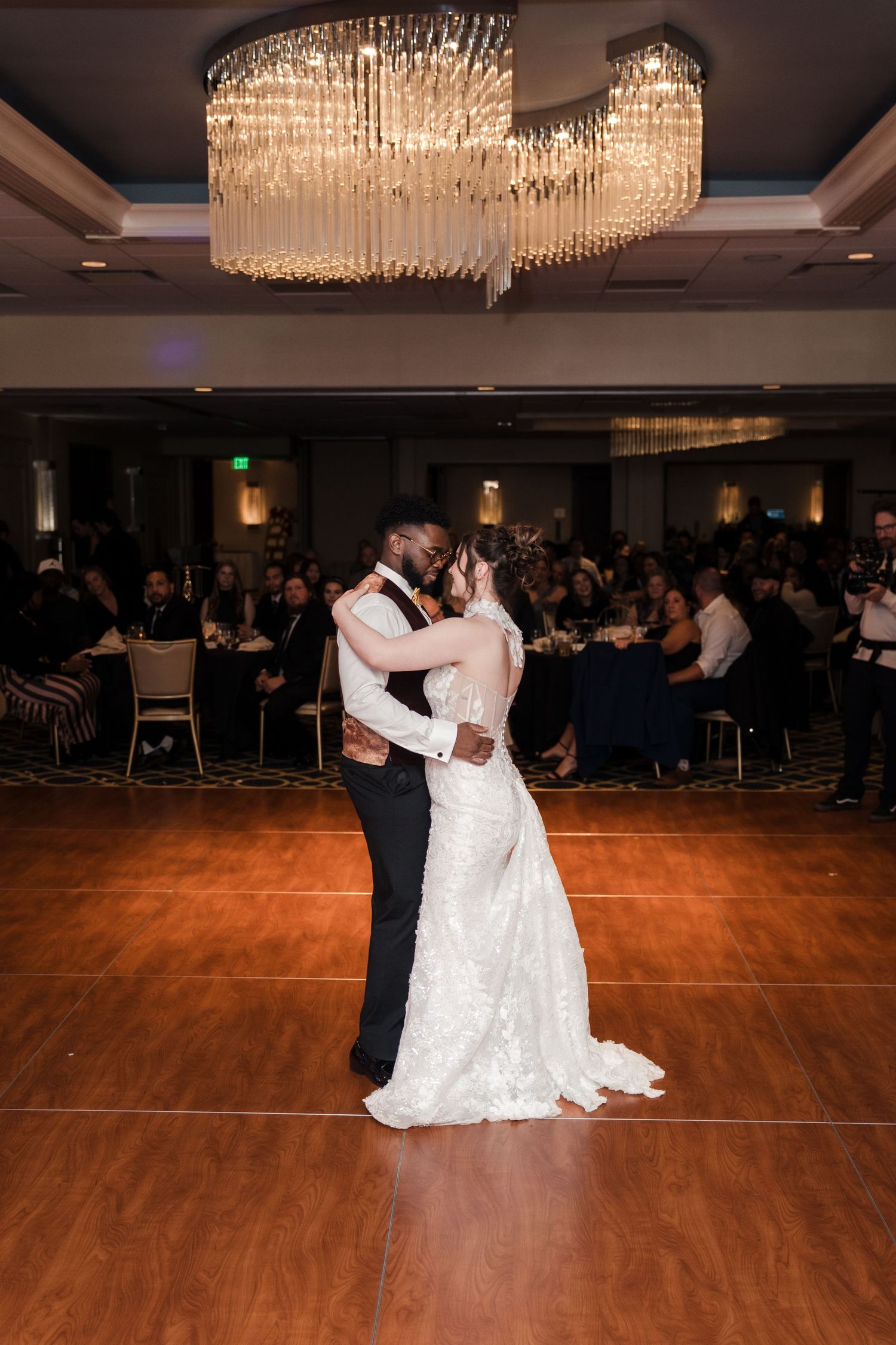 A couple shares a romantic dance on a wooden floor under crystal chandeliers at an elegant wedding reception.