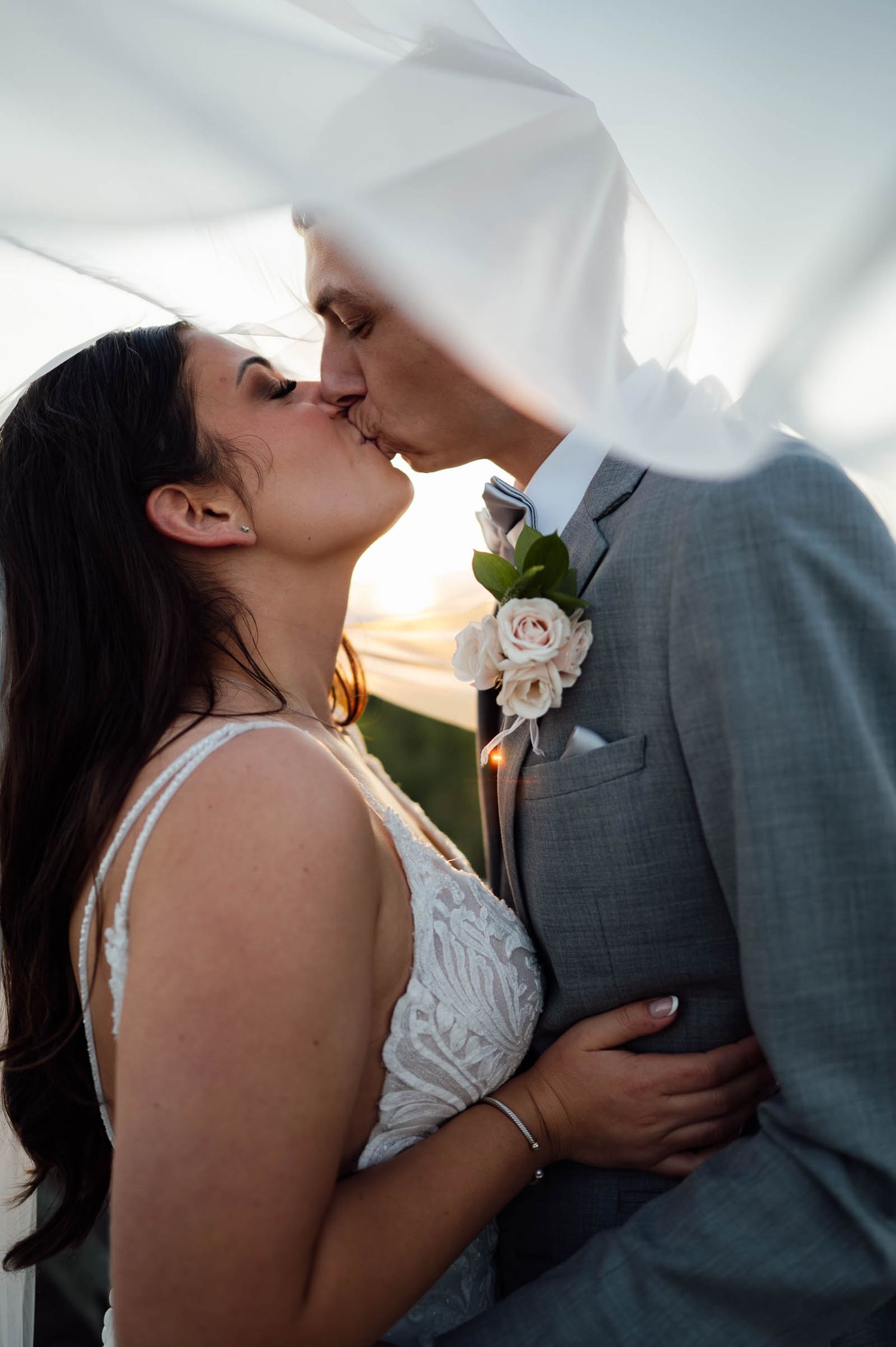 Bride and groom share a romantic kiss under a flowing veil at sunset on their wedding day.